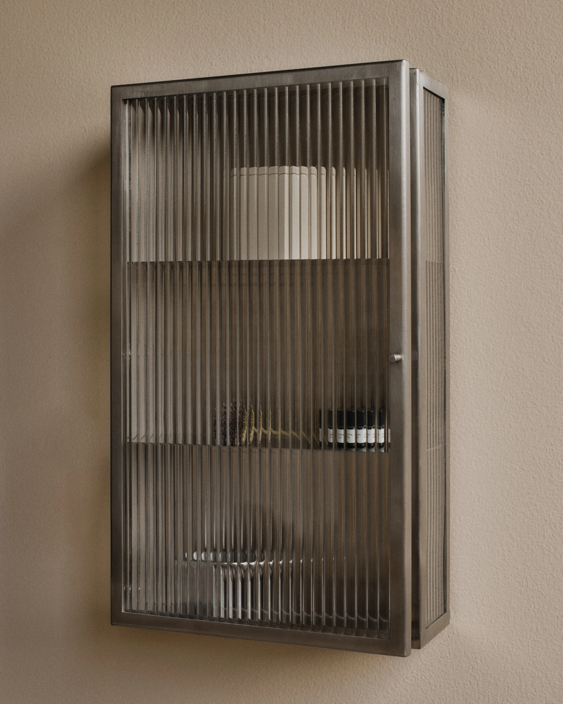 Armoire murale métallique avec portes en verre cannelé, contenant des objets décoratifs et de petites bouteilles.