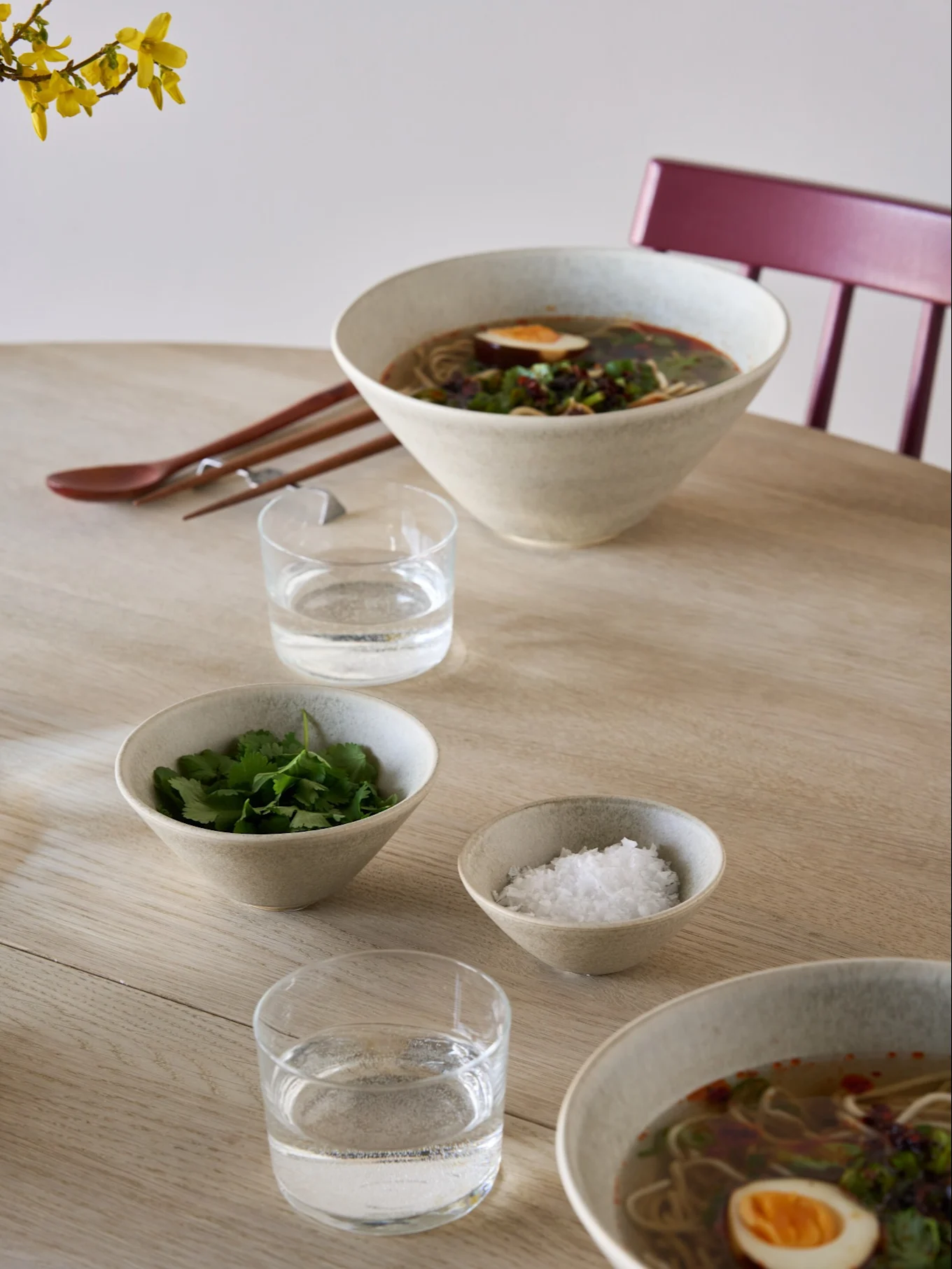 Deux bols de ramen avec œufs et herbes, bols d'accompagnement de coriandre et de sel, eau, baguettes et une cuillère sur une table en bois clair.