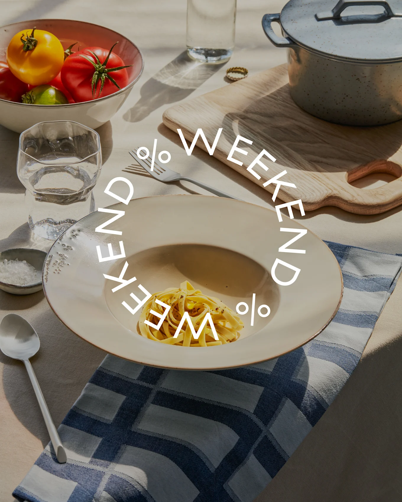 Table avec des pâtes, un bol de tomates fraîches, un verre d'eau et une marmite. Le texte 'WEEKEND % WEEKEND %' est superposé.