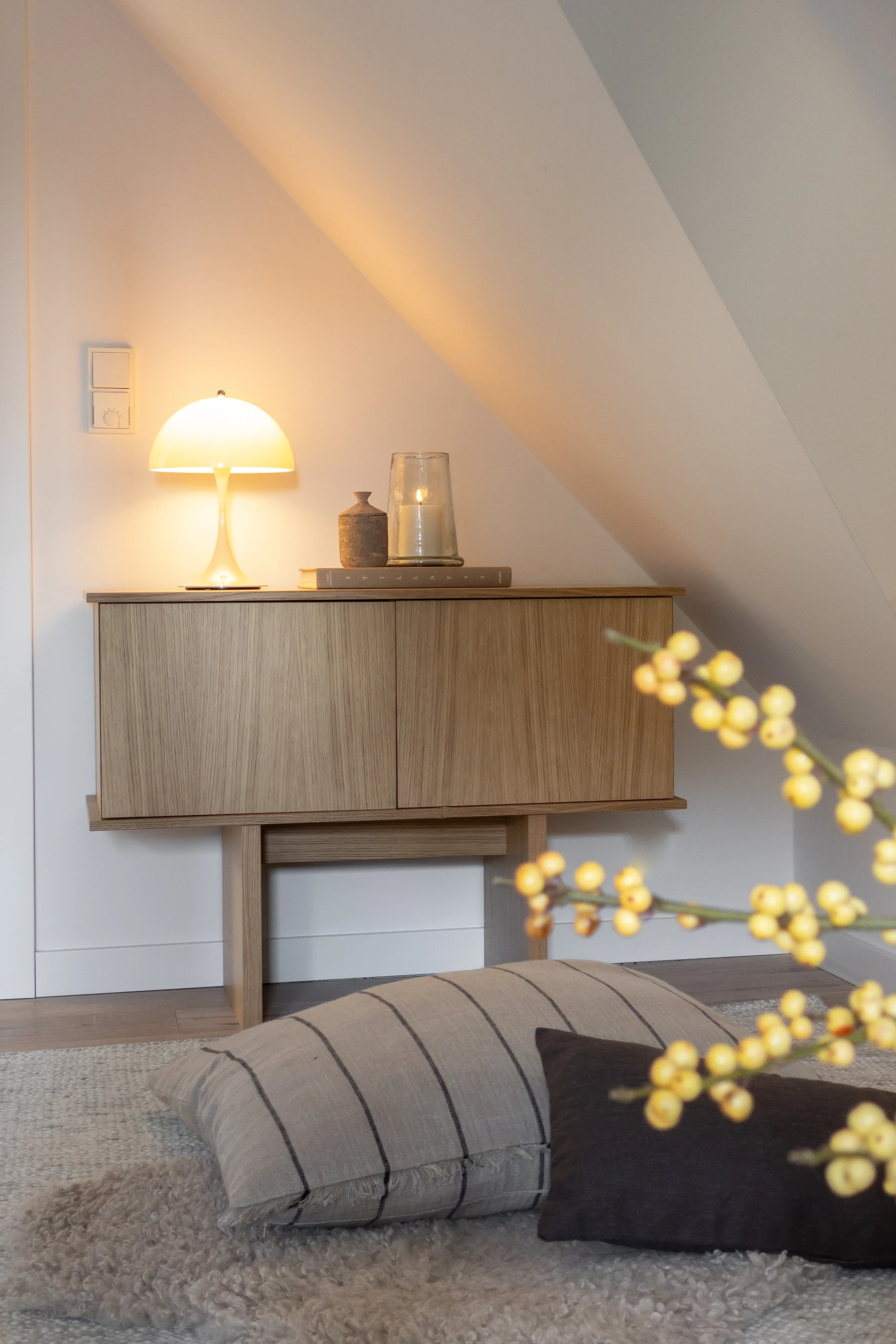 Un meuble en bois avec une lampe champignon lumineuse et une bougie allumée, sur un mur blanc et incliné dans une pièce douillette.