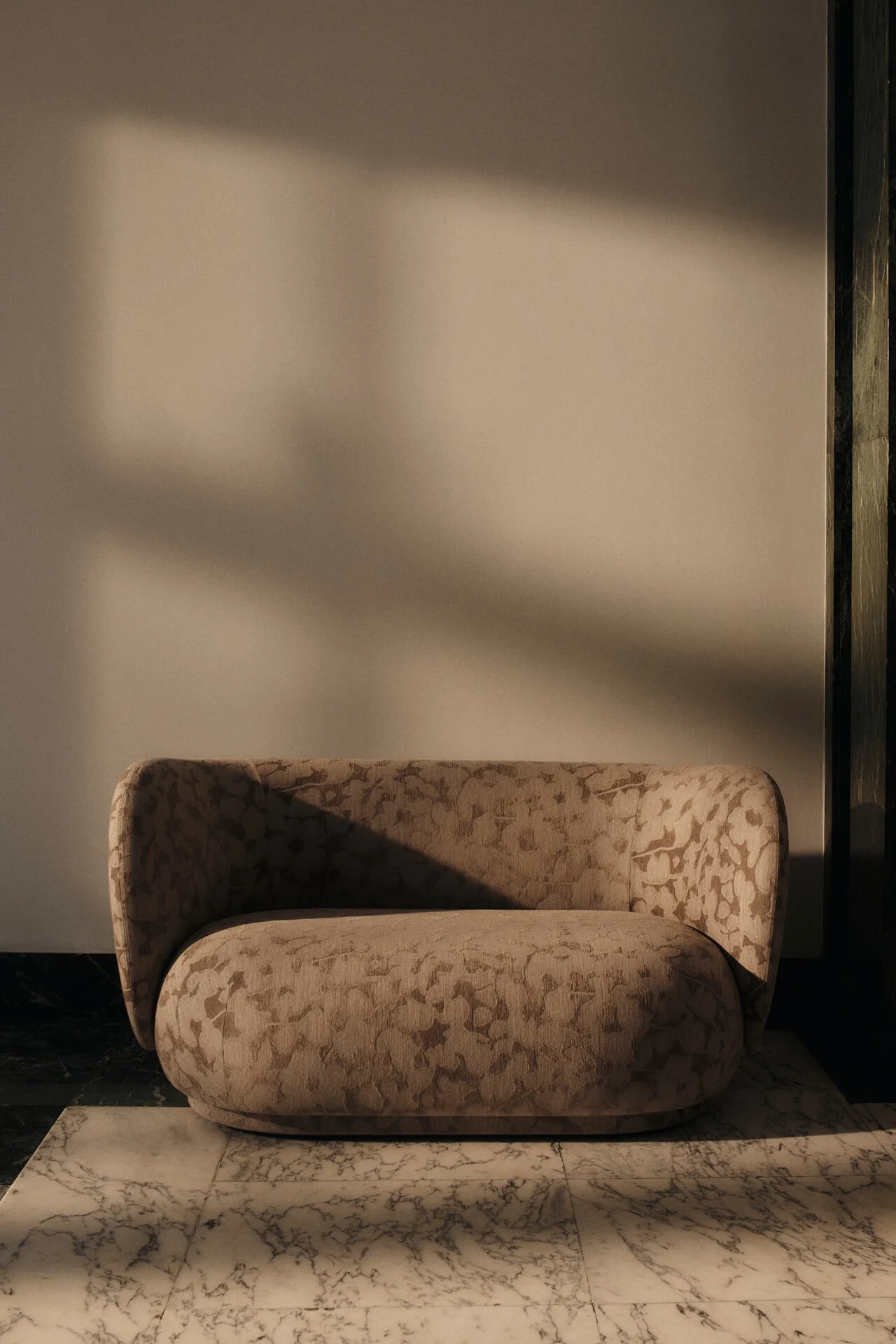 Fauteuil beige arrondi au tissu subtilement motif sur sol en marbre blanc, avec ombres dramatiques de lumière chaude.
