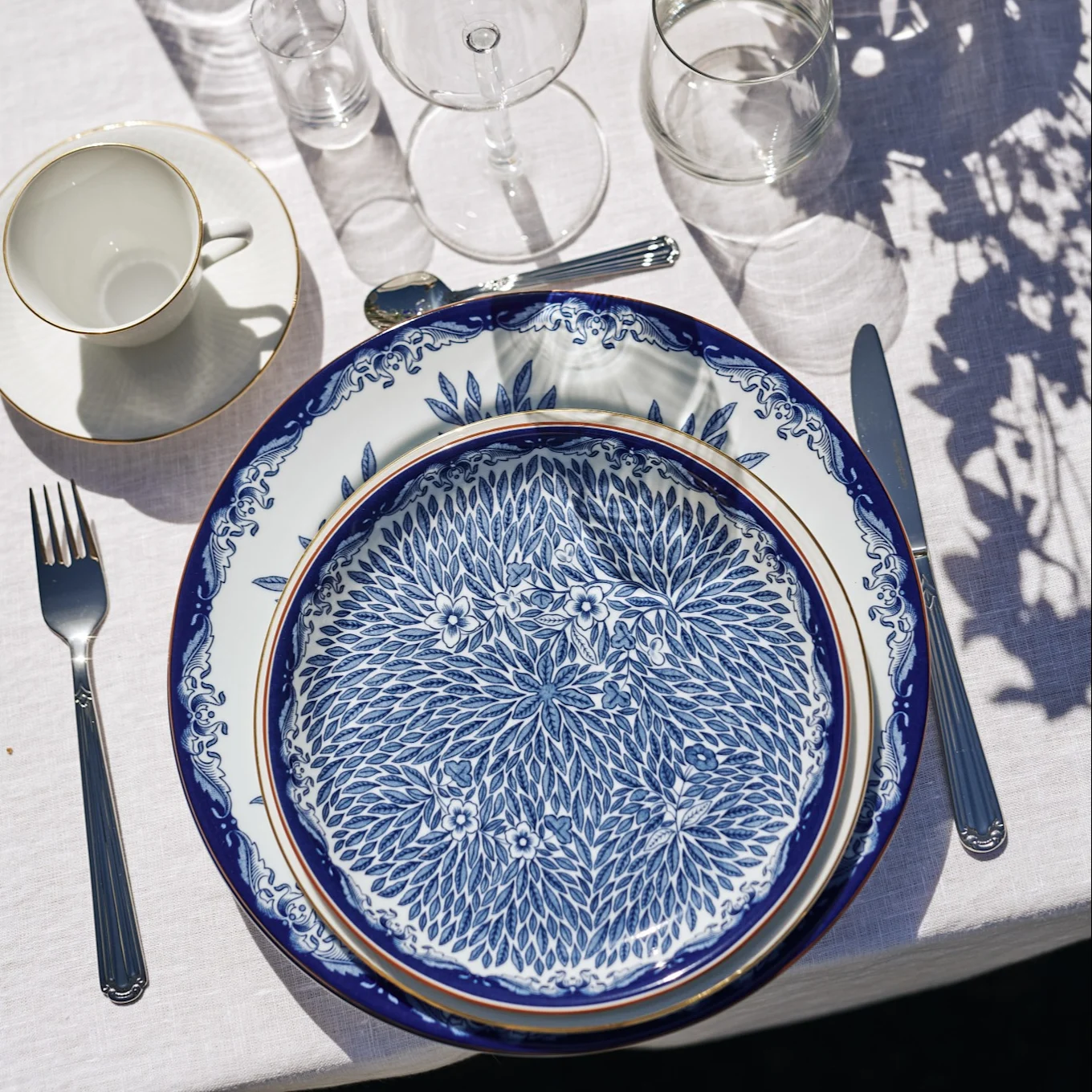 Une table dressée avec des assiettes bleues et blanches à motifs, des couverts, une tasse à thé et des verres sur une nappe blanche sous le soleil.