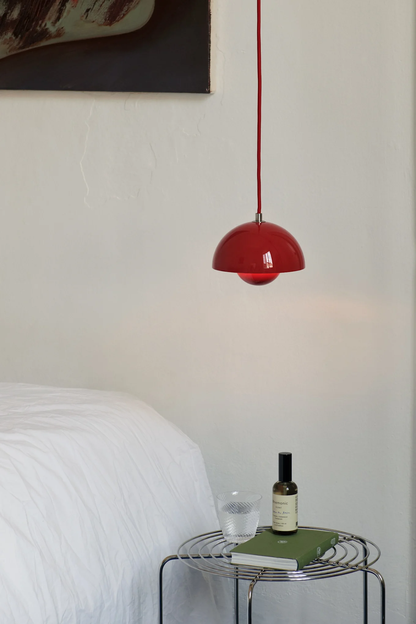 Suspension rouge au-dessus d'une table d'appoint en fil chromé avec un verre d'eau, un livre vert et un flacon pulvérisateur dans une chambre.