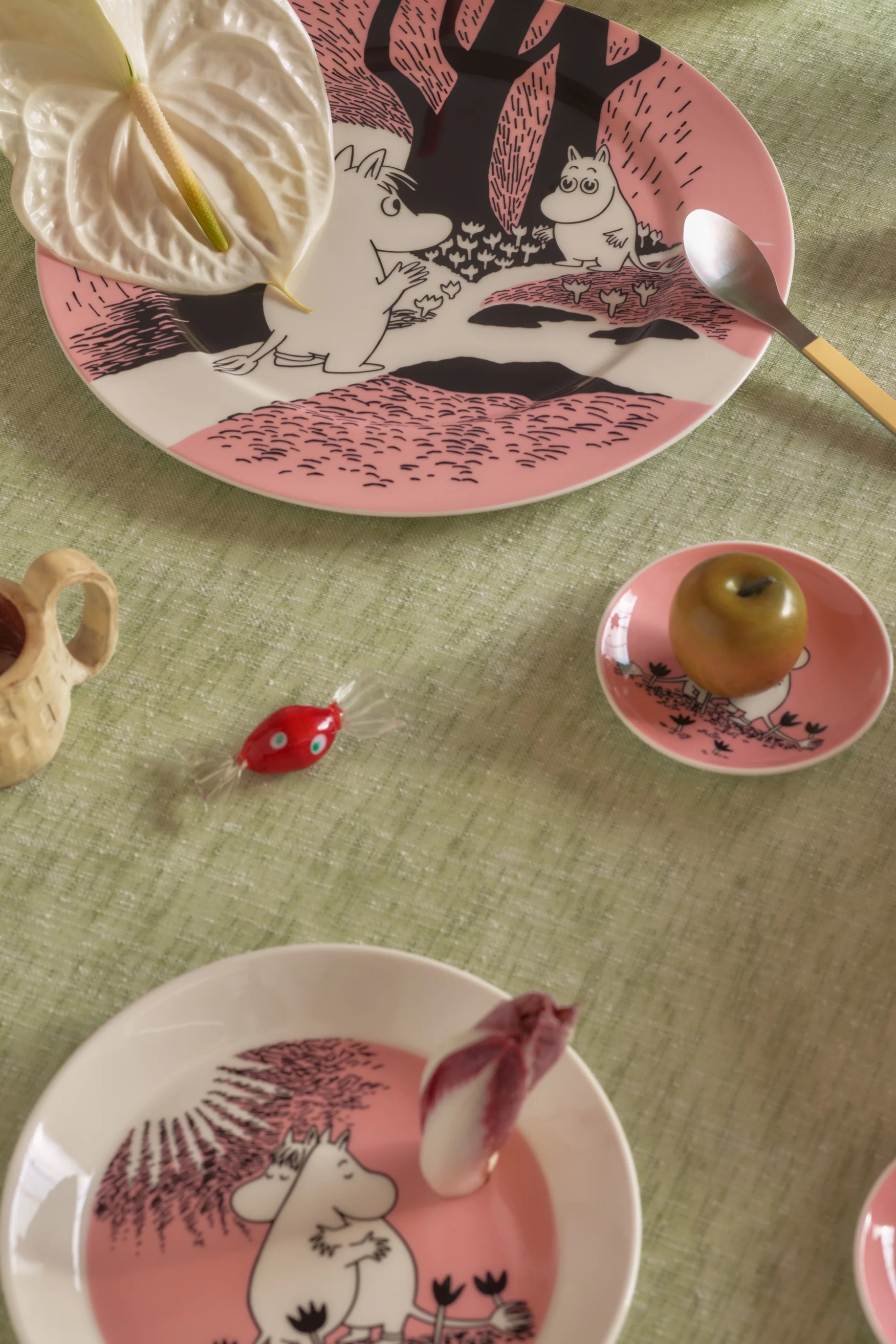 Table dressée avec des assiettes Moomin roses, une fleur d'anthurium, une cuillère, une pomme verte, un bonbon rouge et une tasse en bois sur une nappe verte.