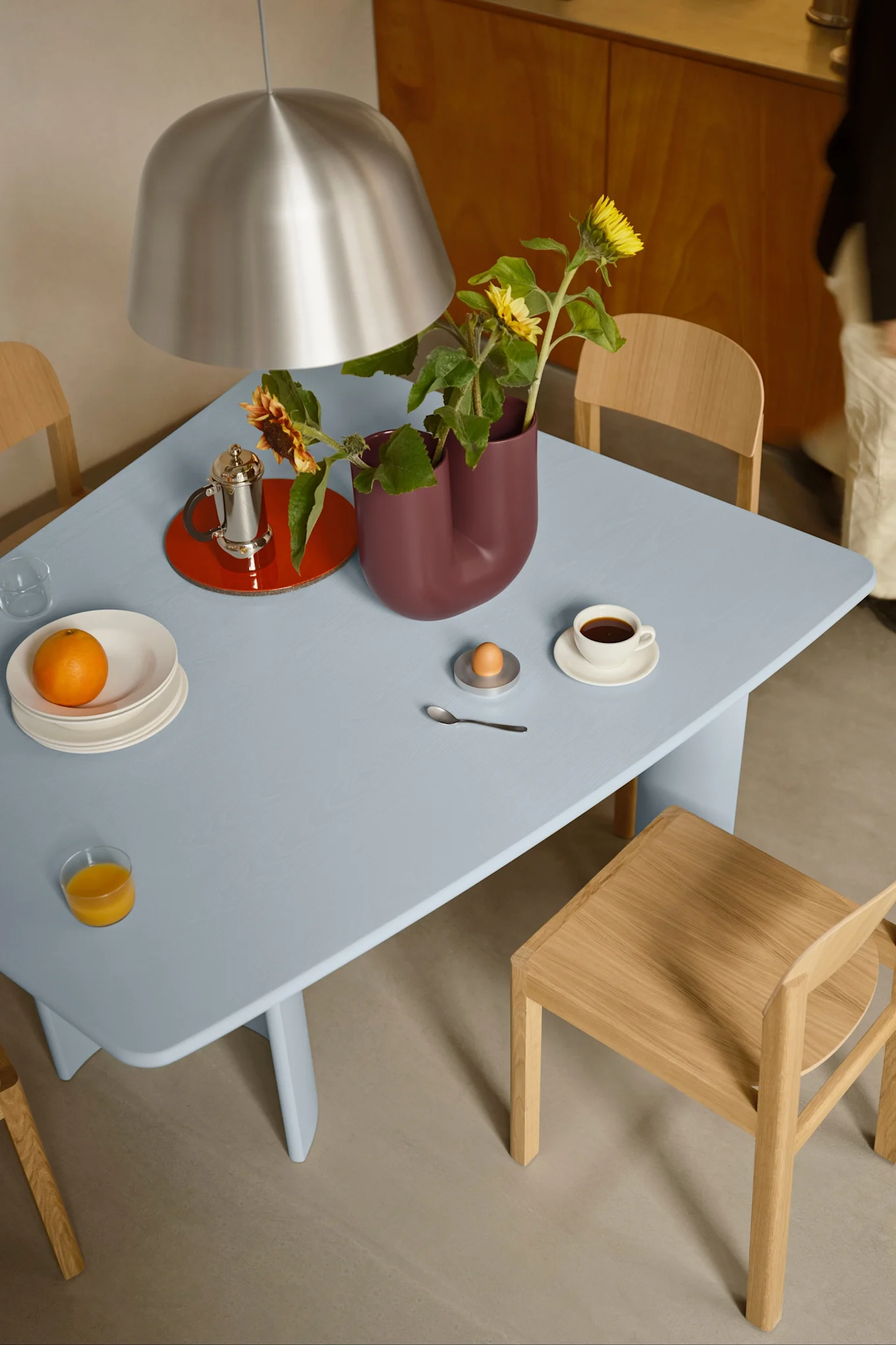 Une table à manger bleue dressée pour le petit-déjeuner, avec du café, du jus d'orange, un œuf et des tournesols dans un vase bordeaux, sous une lampe en argent.