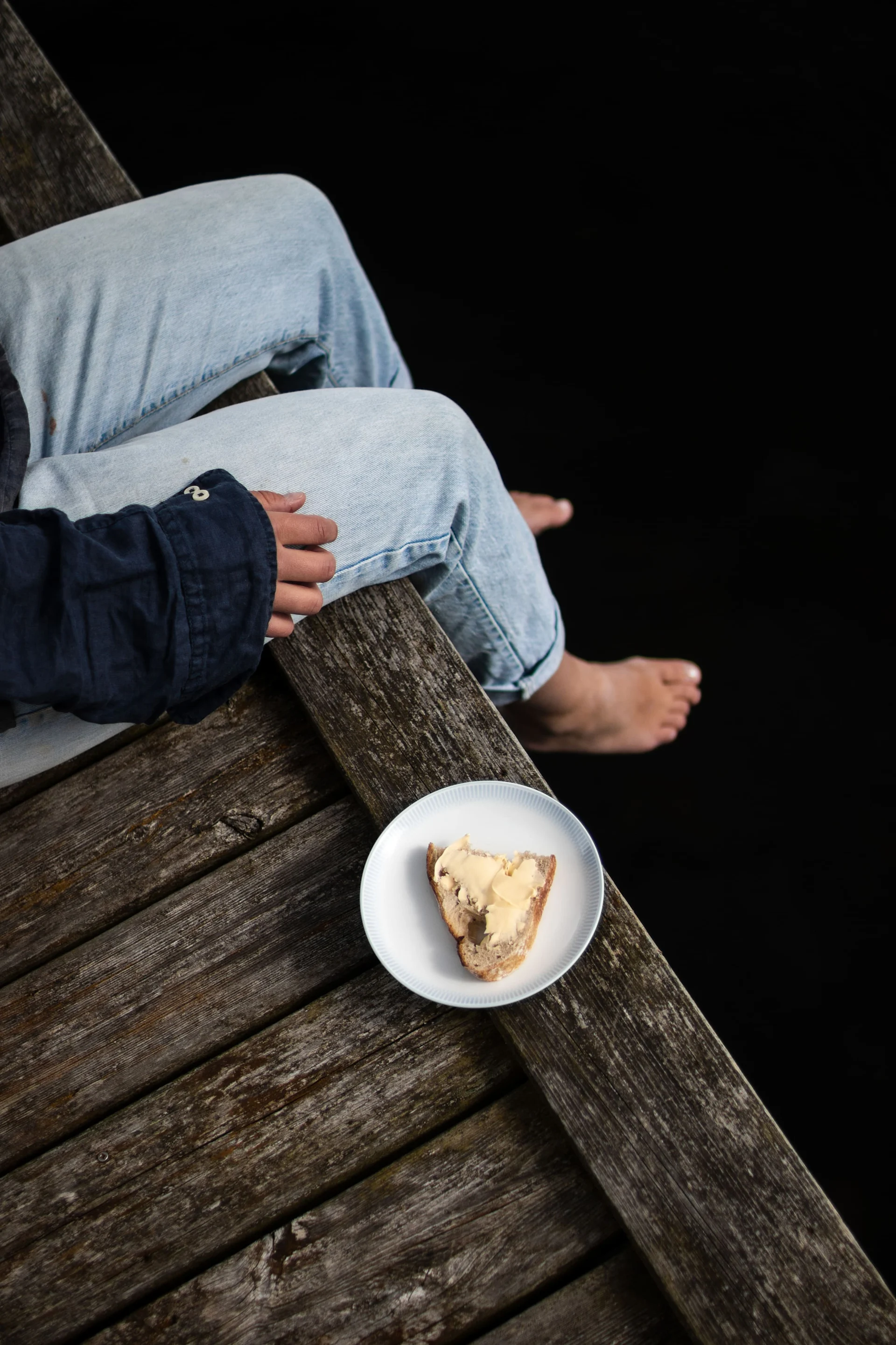 Lagom : une femme assise sur un ponton, à côté d'une assiette Osean de Wik&Walsoe avec une tartine beurrée.