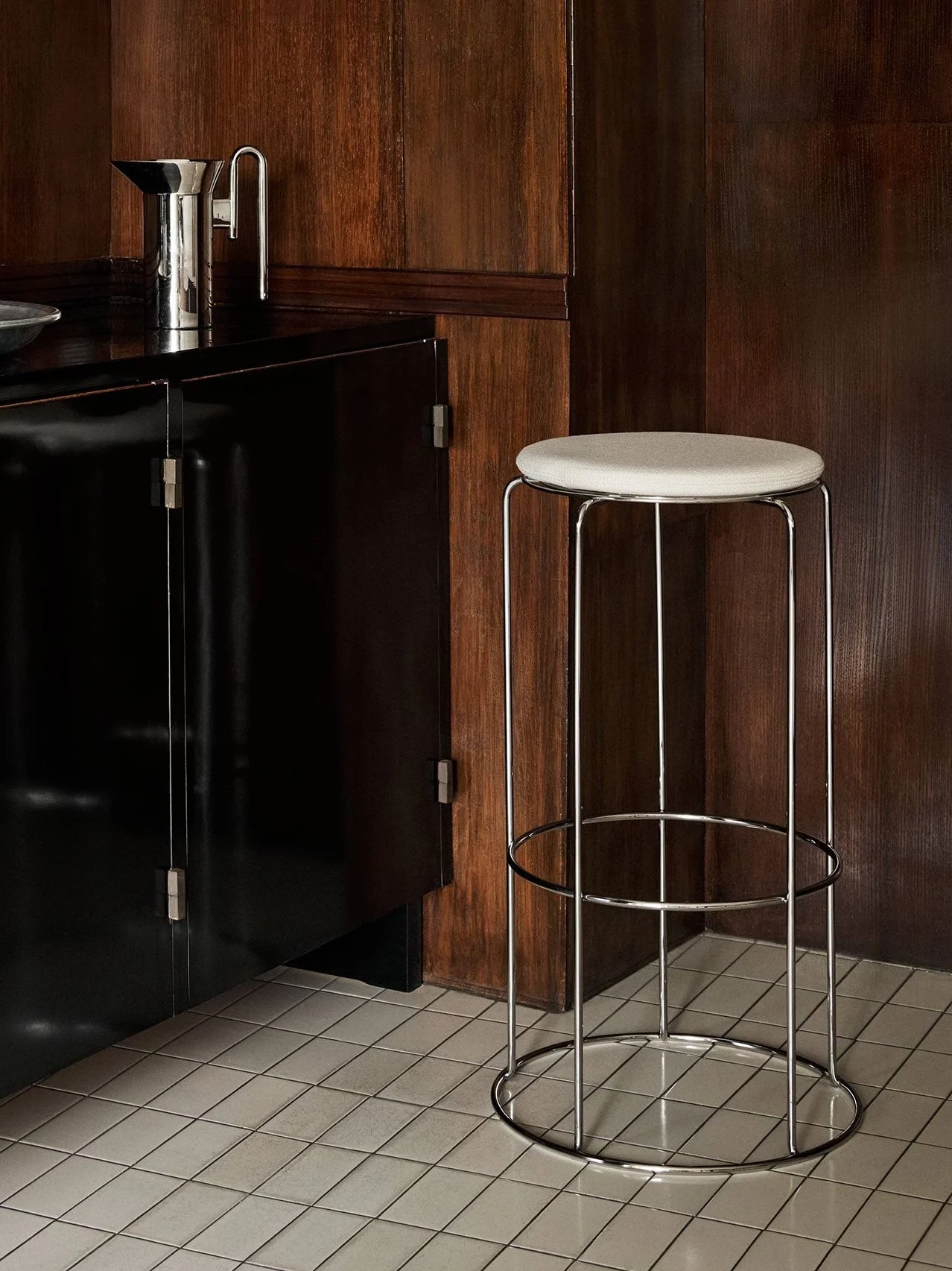 Un meuble noir avec des articles de bar et un tabouret de bar chromé avec une assise claire, devant un revêtement mural en bois foncé et un sol carrelé.
