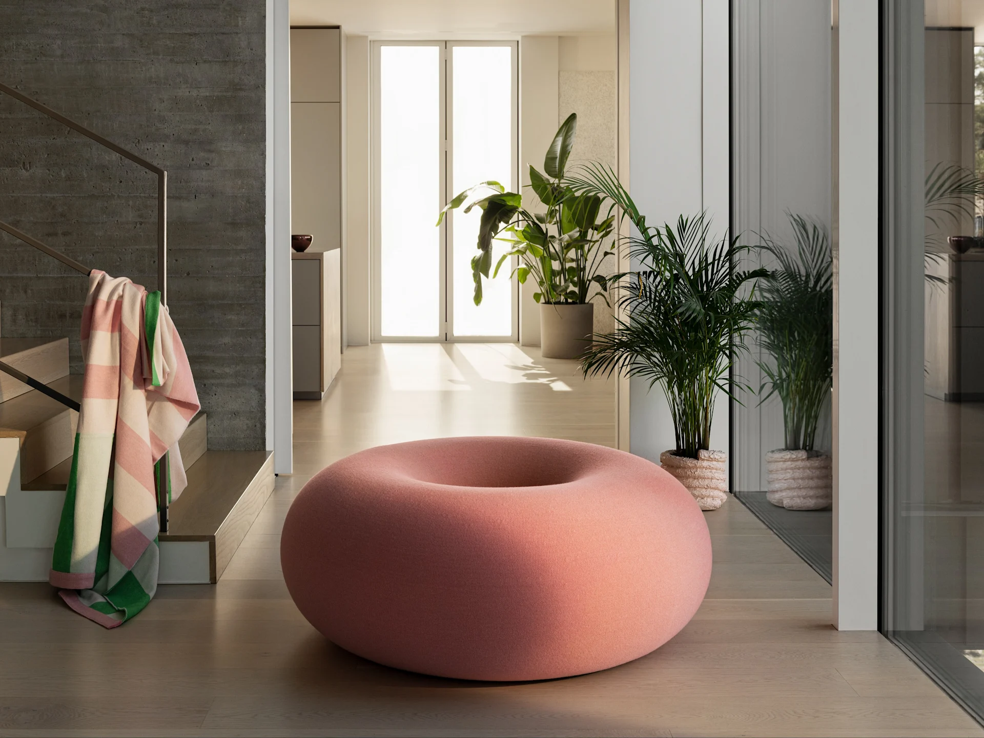 Un grand pouf rose en forme de donut sur un sol en bois clair dans une pièce moderne avec des plantes vertes et un mur en béton.