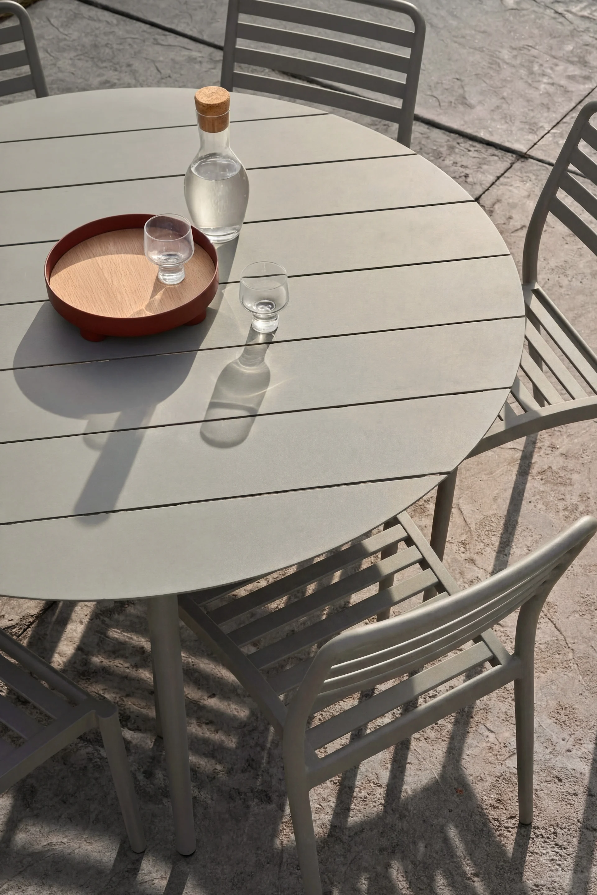 Une table d'extérieur ronde grise, trois chaises. Une carafe en verre remplie d'eau et deux verres, dont l'un posé sur un plateau en bois, sont posés sur la table.