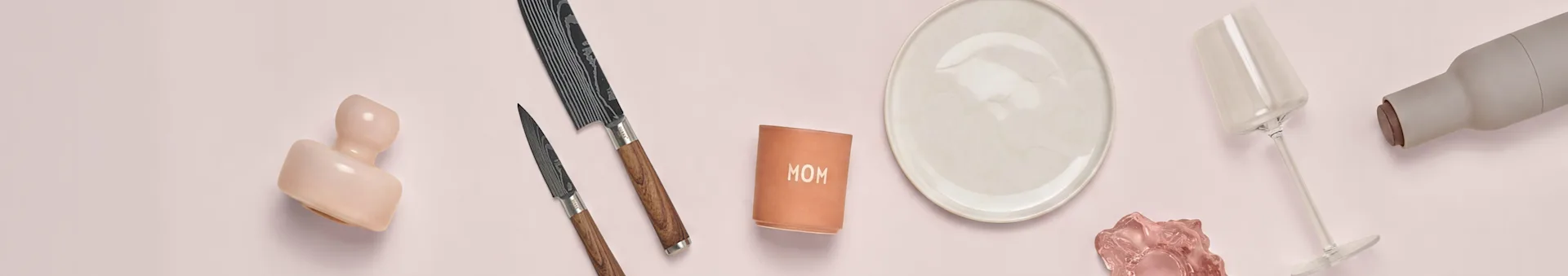 Céramique rose, deux couteaux, tasse 'MOM', assiette blanche, verre rose, verre à vin, bouteille grise sur fond rose.