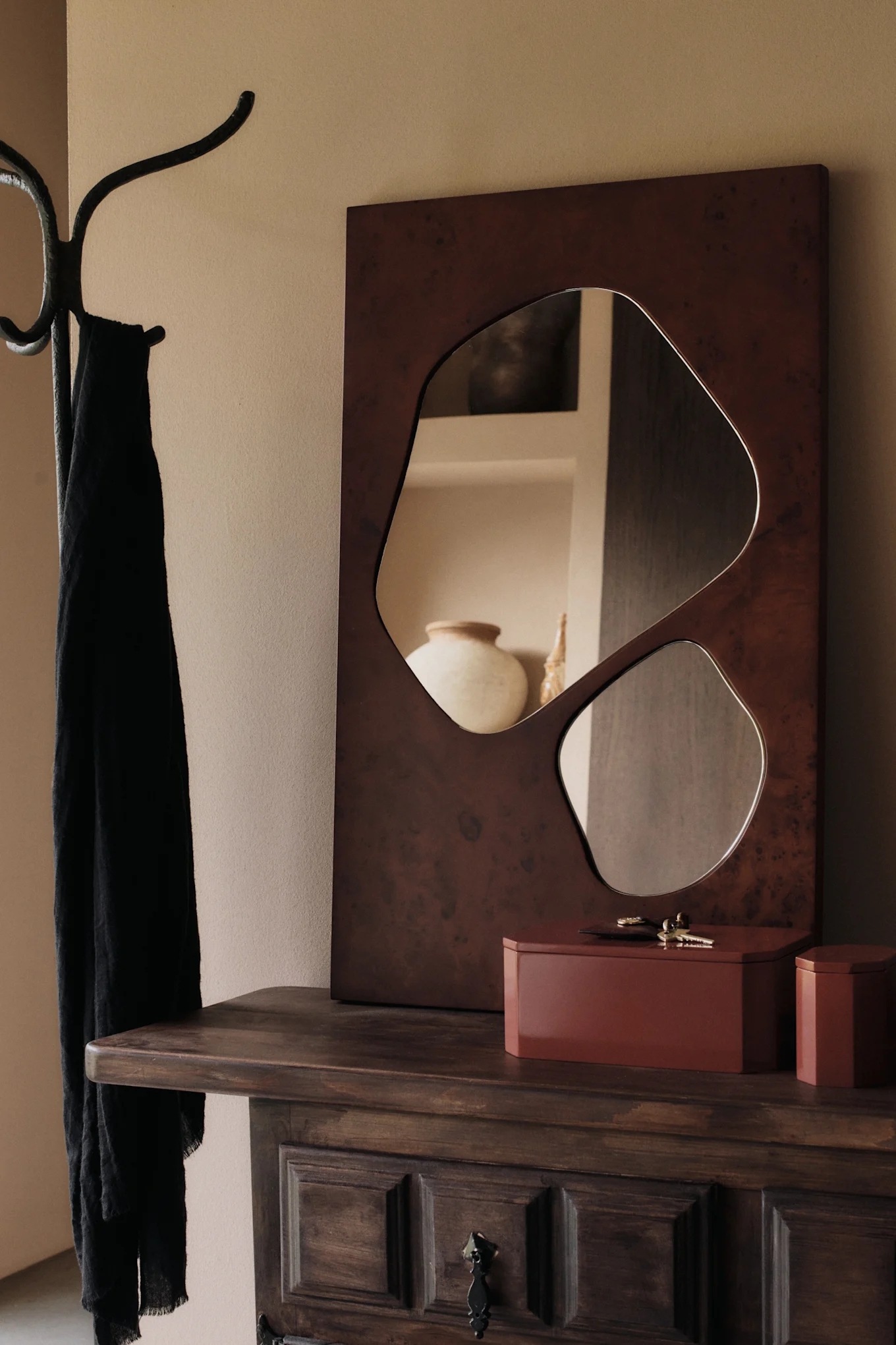 Un miroir de forme organique et deux boîtes brun-rouge sur une commode en bois à côté d'un porte-manteau avec une écharpe noire.