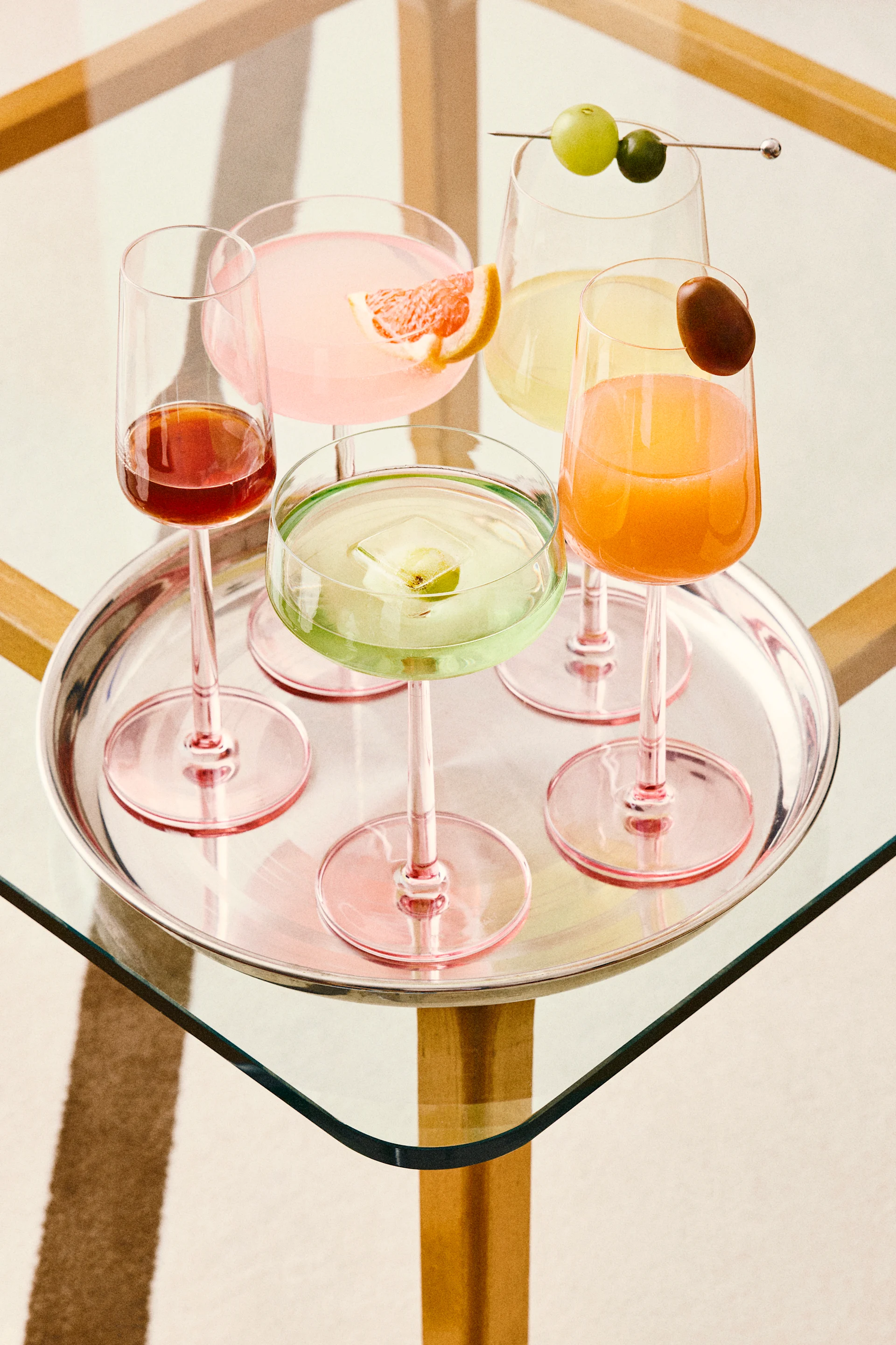 Cinq cocktails colorés sur un plateau en argent, y compris des boissons roses, vertes, oranges et rouges avec diverses garnitures, sur une table en verre.