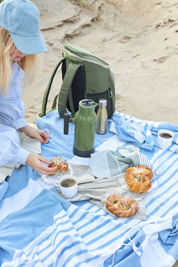 Ici, vous voyez une femme assise sur une couverture de pique-nique à rayures bleues avec un sac de pique-nique vert, le tout provenant de Sagaform.