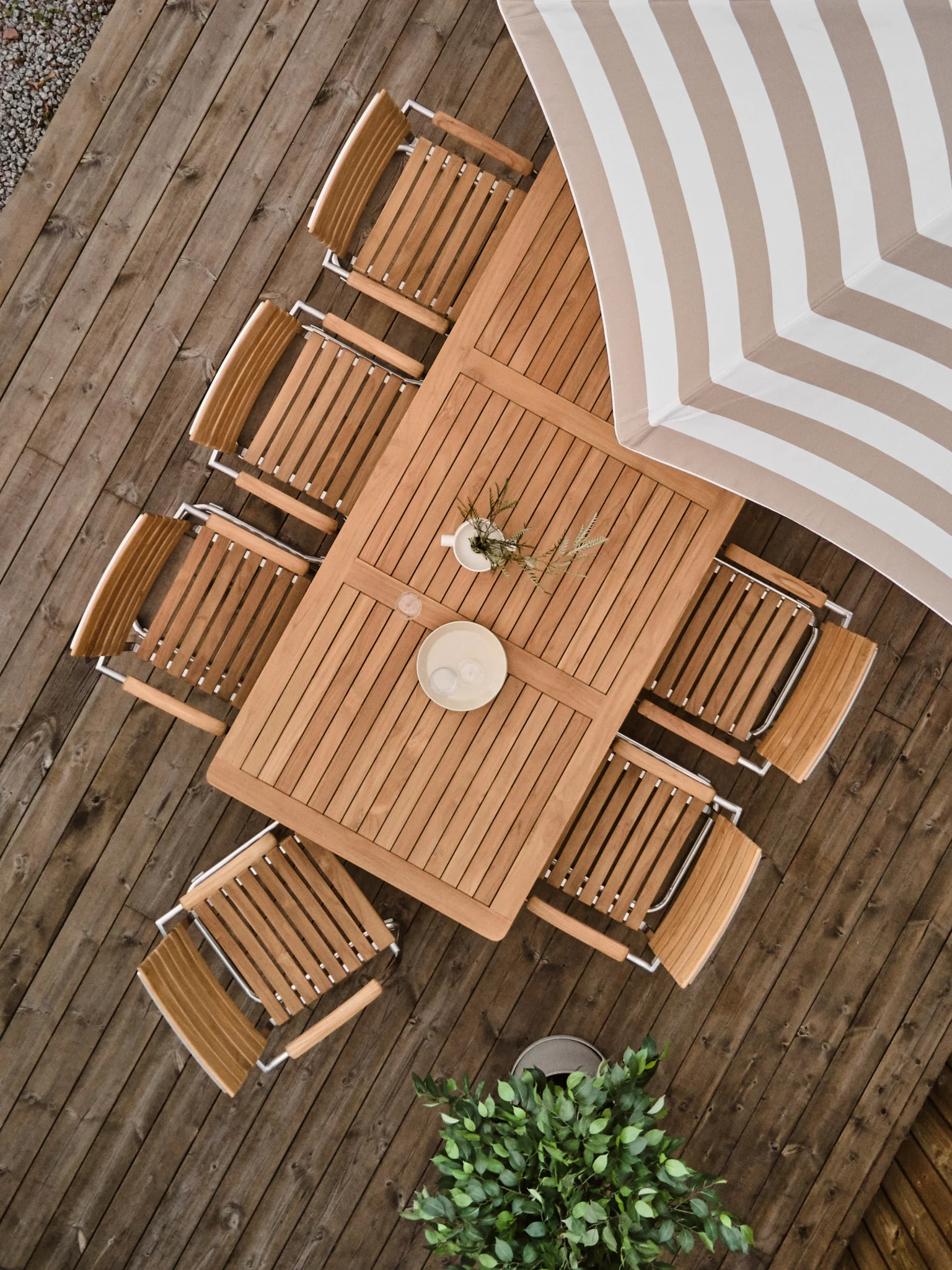 Vue plongeante d'un ensemble de salle à manger d'extérieur en bois sur une terrasse, avec une table rectangulaire, huit chaises et un parasol rayé.