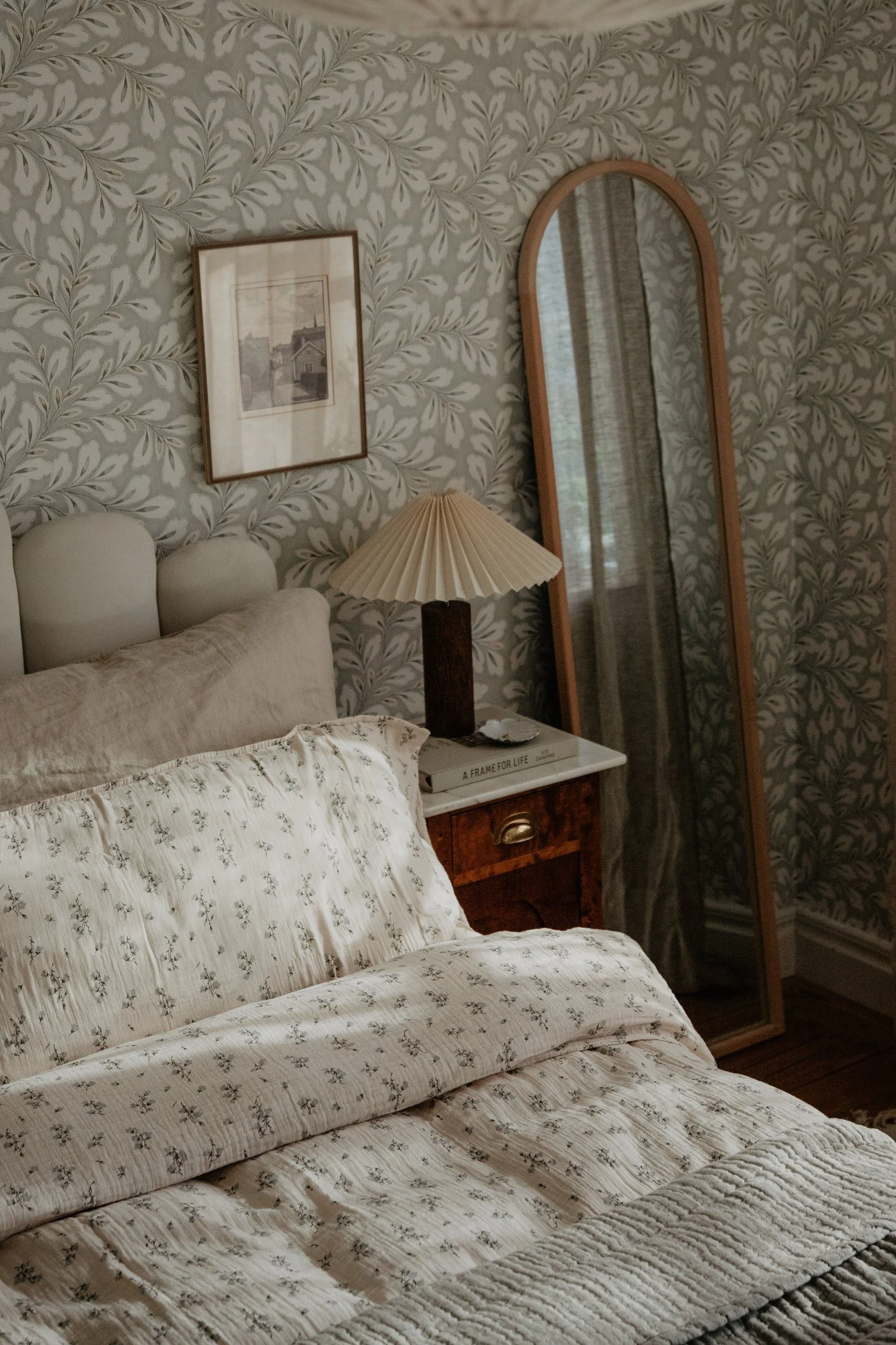 Une chambre à coucher avec une parure de lit à motifs couleur crème, une lampe plissée posée sur une table de chevet en bois et un grand miroir, le tout sur fond de papier peint à motifs de feuillage.