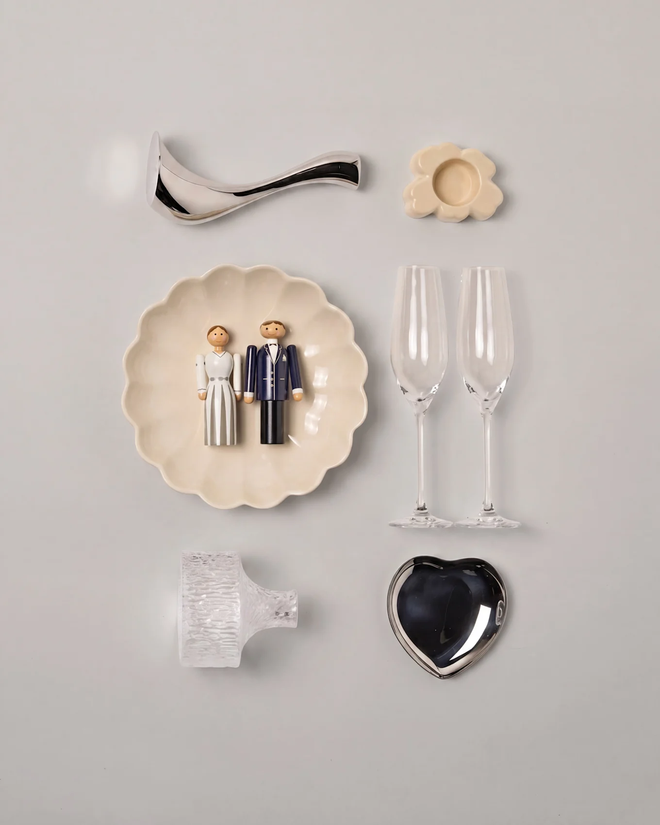 Composition de mariage à plat avec figurines en bois de mariés sur une assiette festonnée, deux flûtes à champagne, un plat cœur argenté et divers objets décoratifs.