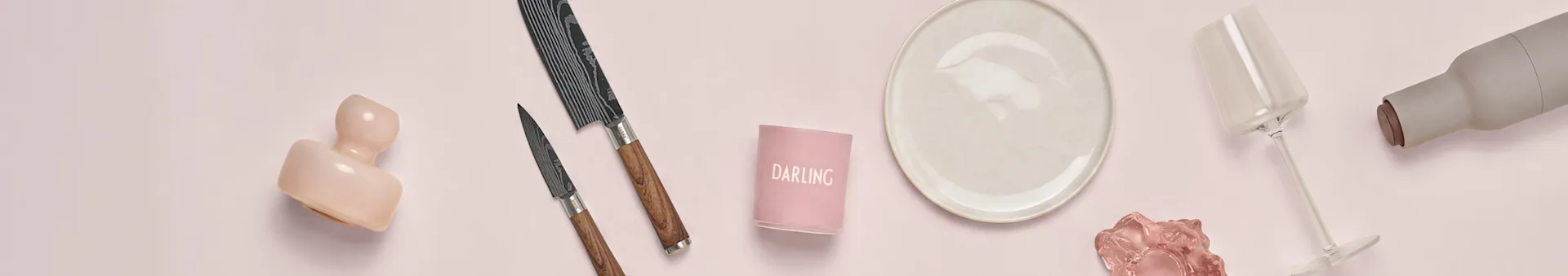 Assortiment d'articles de cuisine et de salle à manger sur fond rose : une sculpture rose, deux couteaux à motifs, un bougeoir "DARLING", une assiette crème, un verre à vin transparent et une bouteille grise.