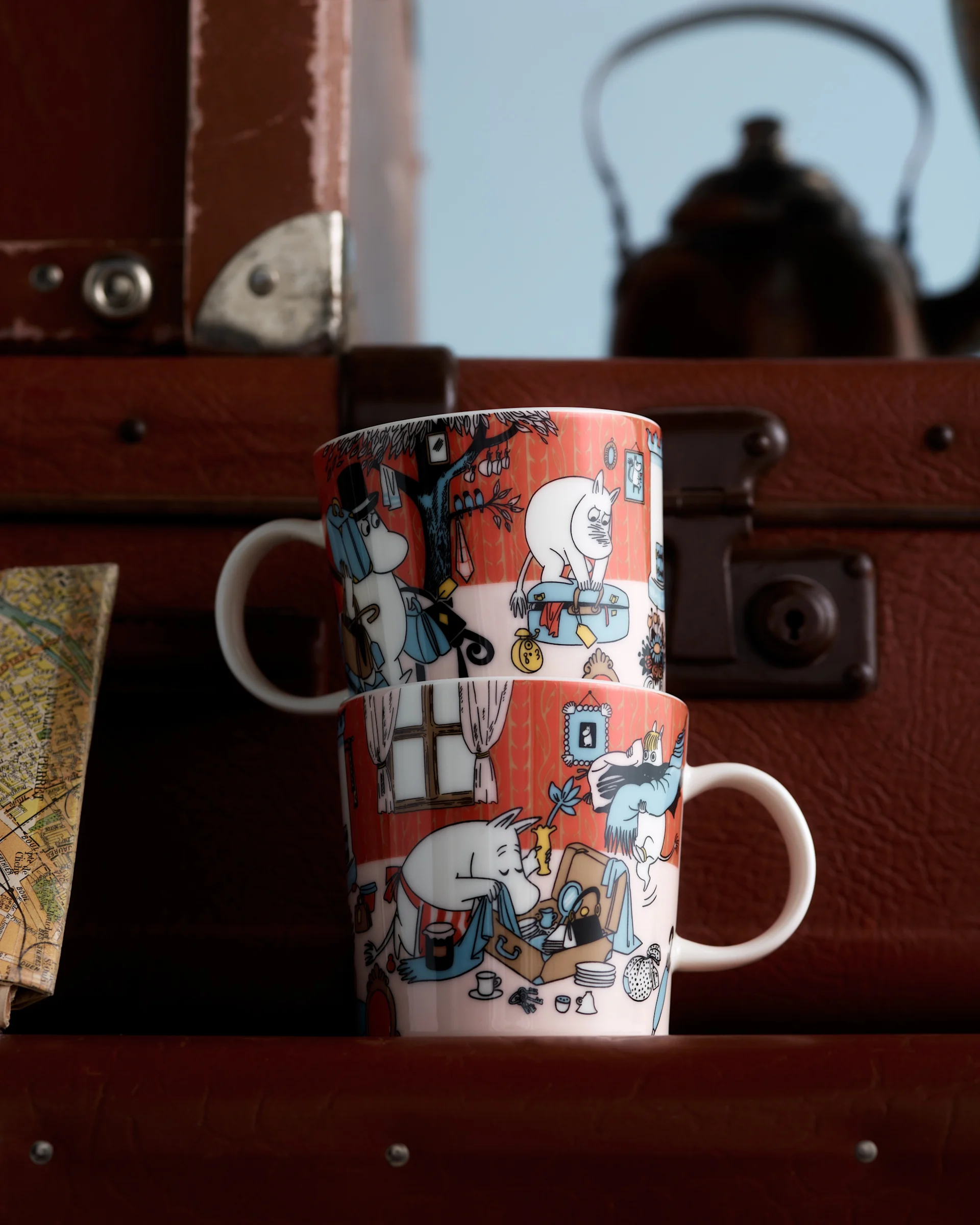 Deux tasses Moomin avec des illustrations de personnages rouge-orangé, empilées devant des valises marron et une carte floue.