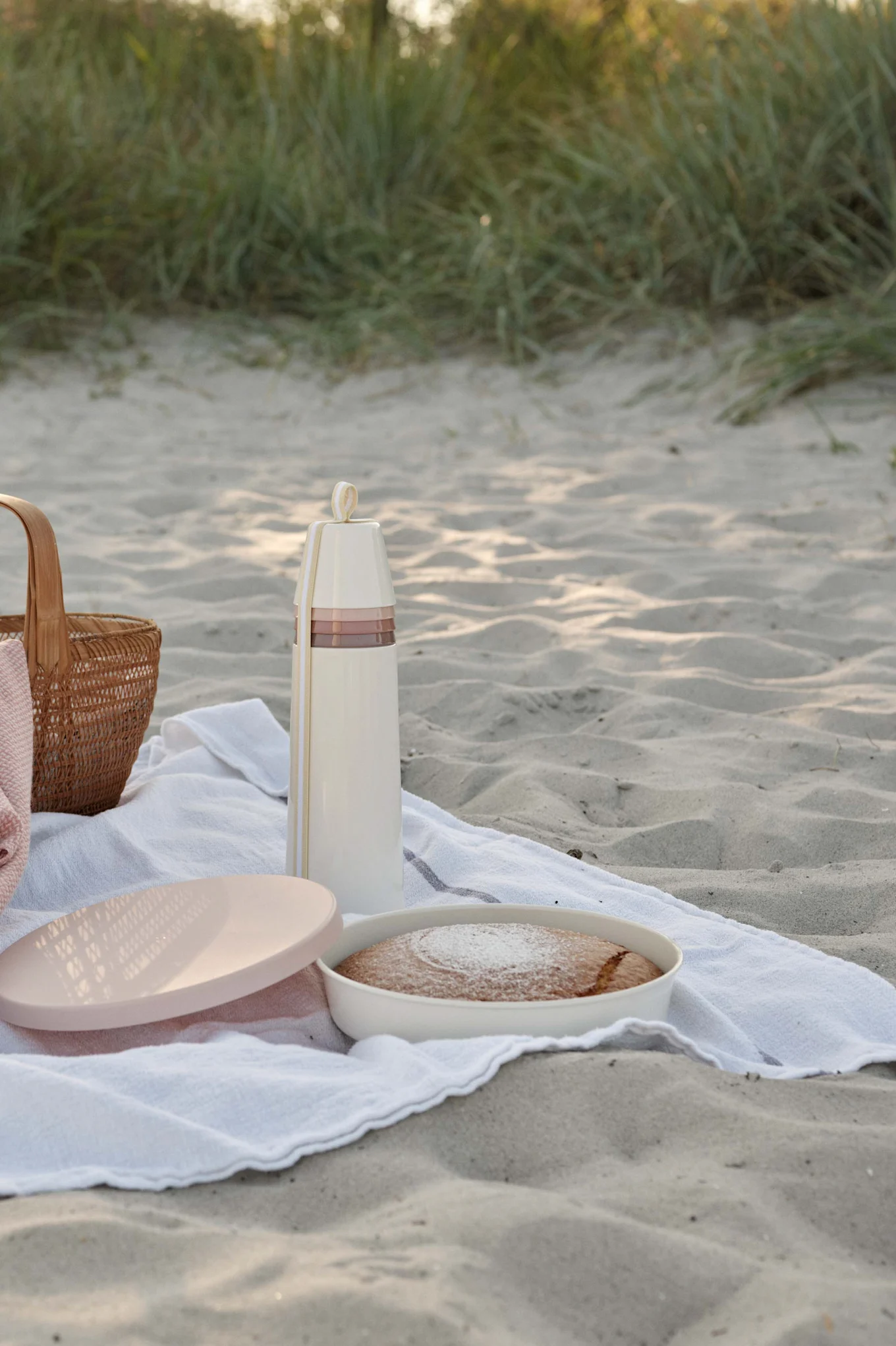 Pique-nique sur une plage de sable avec un thermos couleur crème, un gâteau saupoudré de sucre glace et un panier tressé.