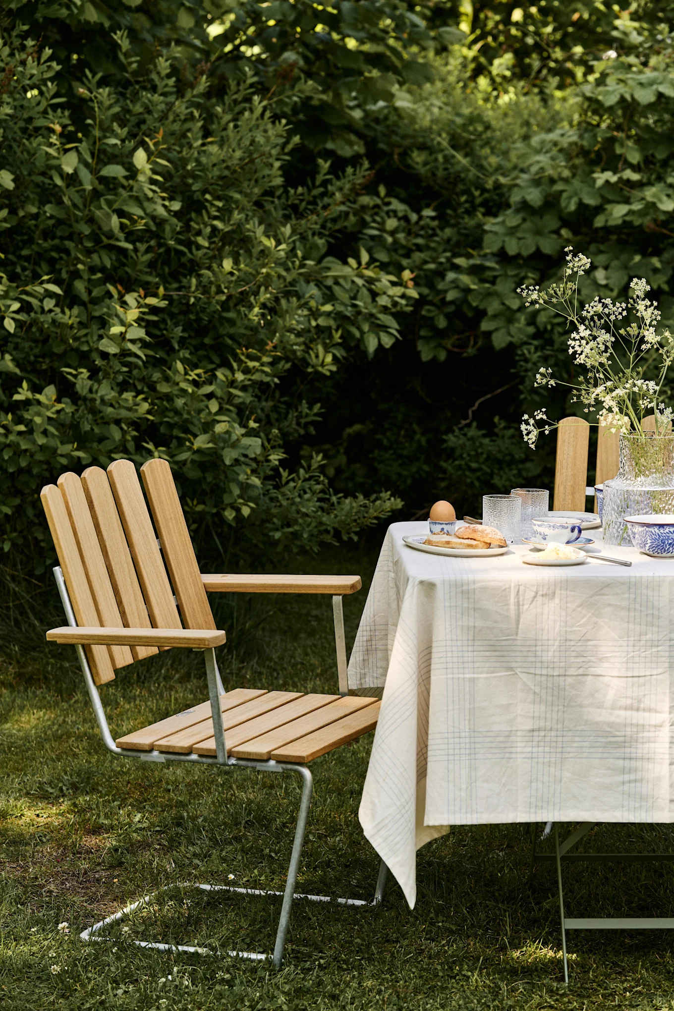 Une scène de petit-déjeuner en plein air, avec une chaise en lattes de bois clair et un dossier en métal, placée à côté d'une table dressée d'une nappe à carreaux, de vaisselle et d'un vase de fleurs blanches.