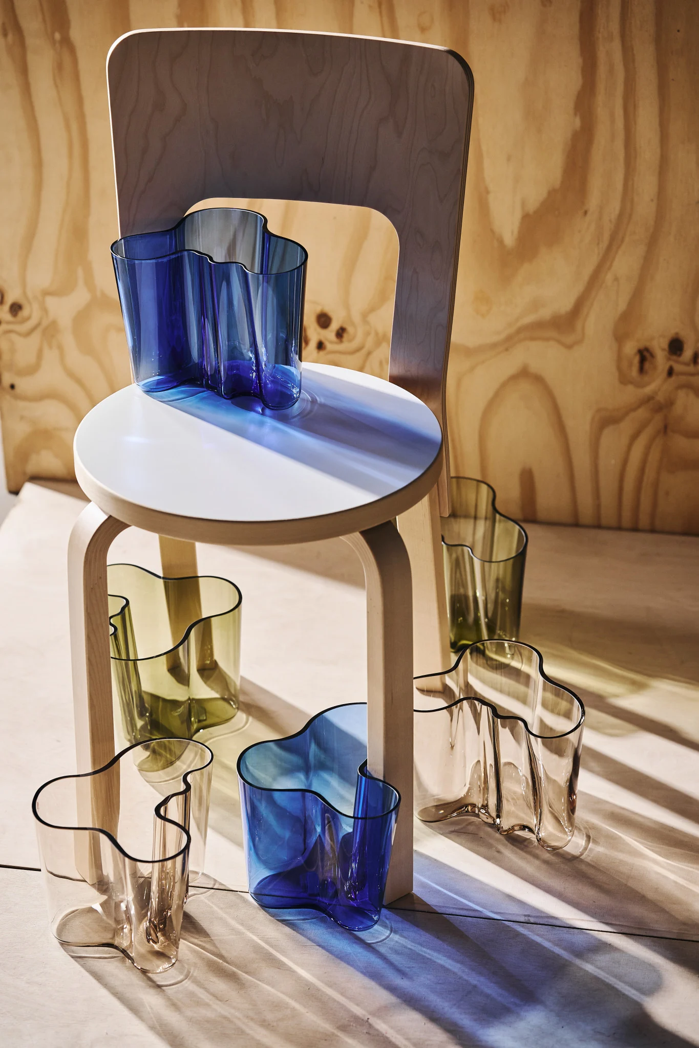 Plusieurs vases en verre ondulés bleus, vert olive et transparents, disposés sur et autour d'un tabouret en bois clair avec une chaise en bois en arrière-plan. La lumière du soleil projette des ombres colorées.