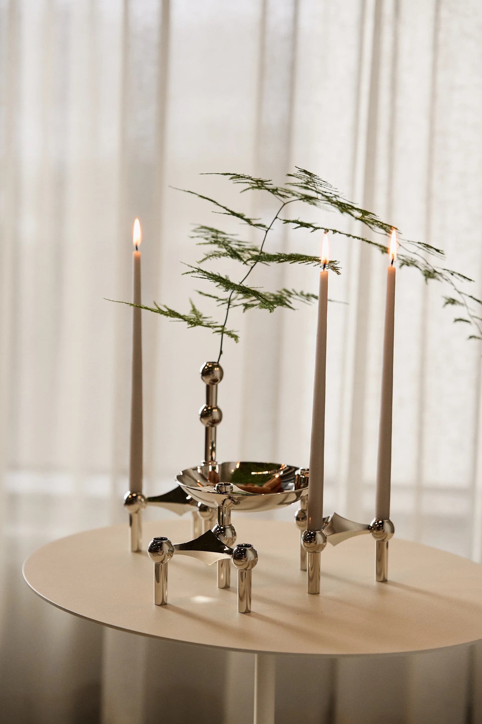 Trois bougies allumées dans un bougeoir modulaire en argent orné de feuillage vert, posées sur une petite table ronde.
