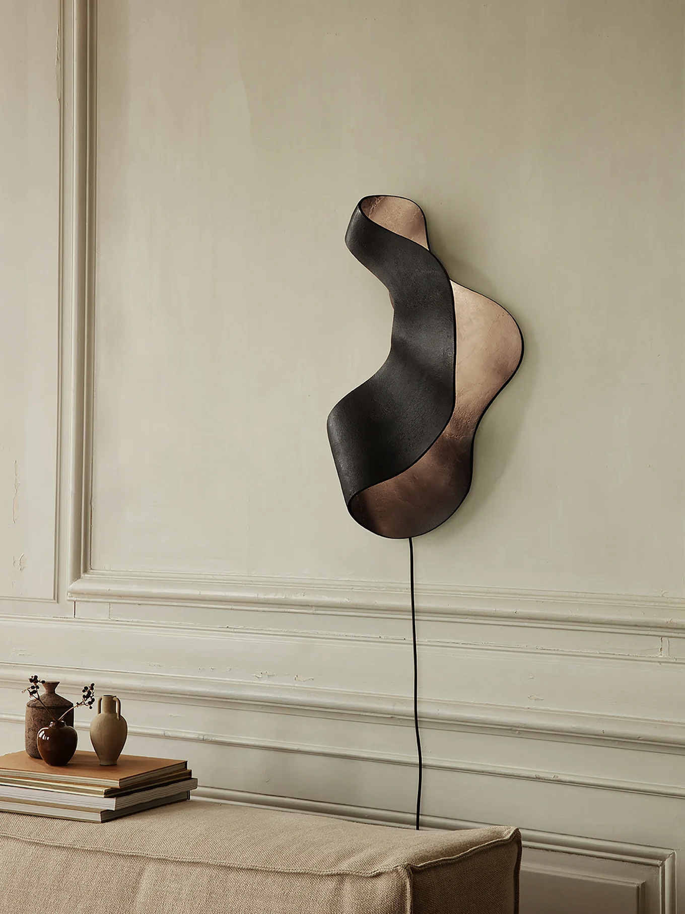 Applique murale ondulée noire et bronze sur mur clair, au-dessus d'un canapé avec vases et livres.