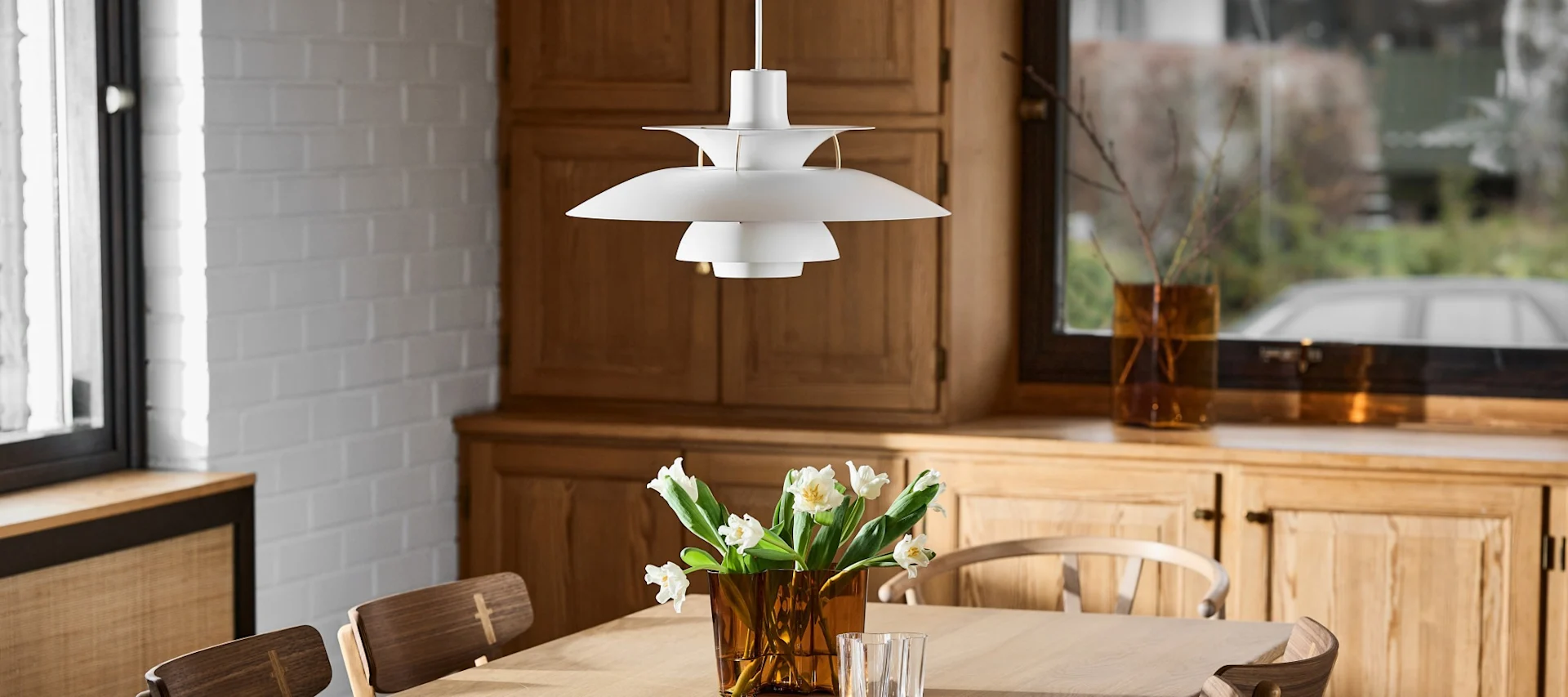 Une lampe suspendue blanche à plusieurs niveaux est au-dessus d'une table en bois avec un vase de tulipes blanches, dans une pièce avec des armoires en bois.