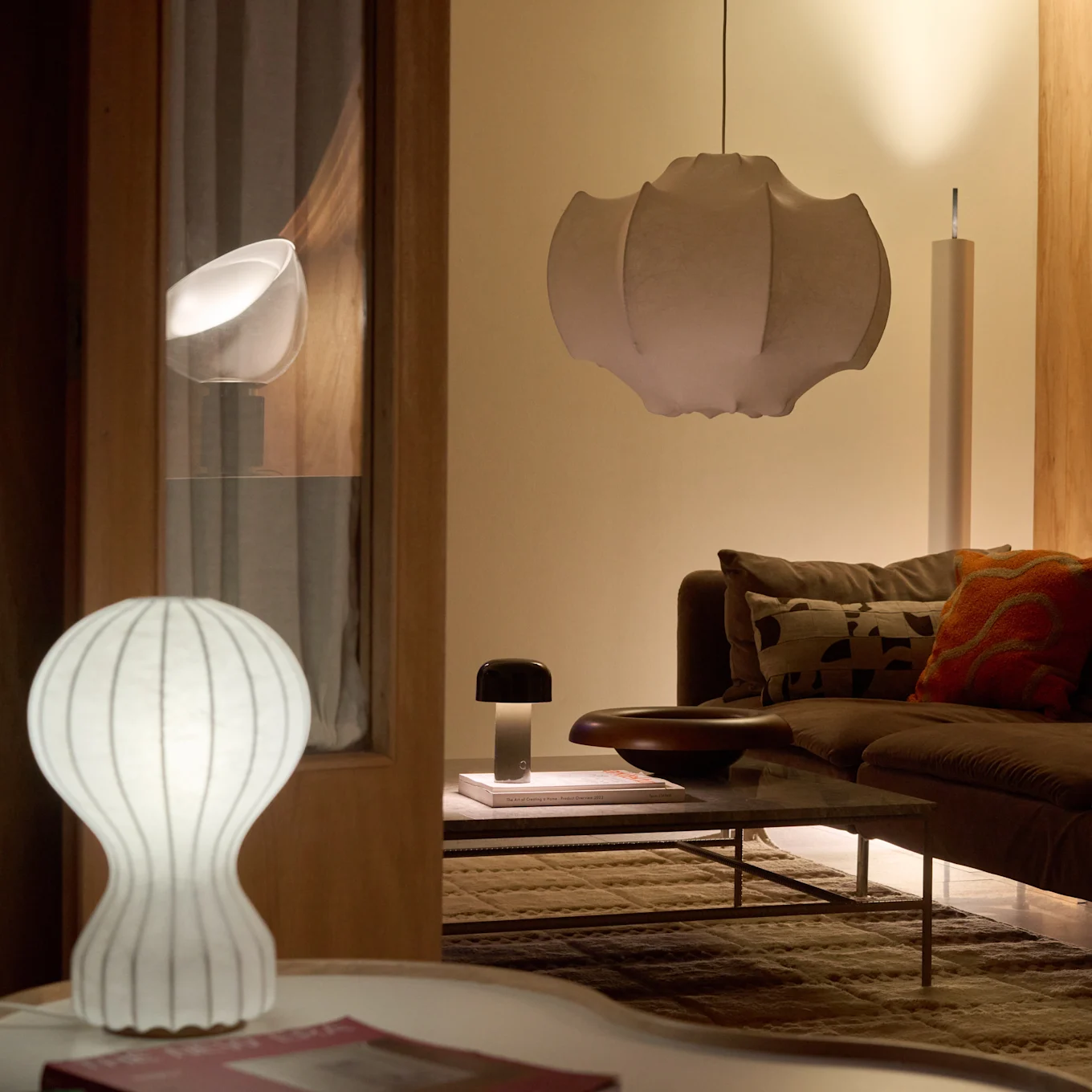 Un salon avec un canapé et une table basse marron, éclairé par une lampe de table blanche côtelée, une lampe champignon foncée et une grande suspension plissée, créant une lueur chaleureuse.