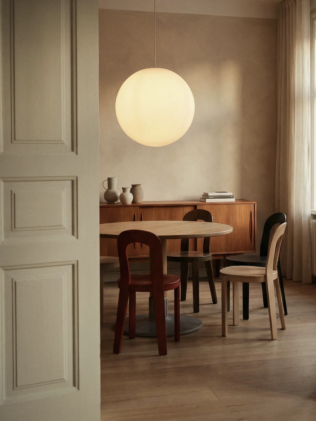 Une salle à manger moderne avec une table ronde en bois, des chaises variées, une grande suspension sphérique et une commode avec des vases.