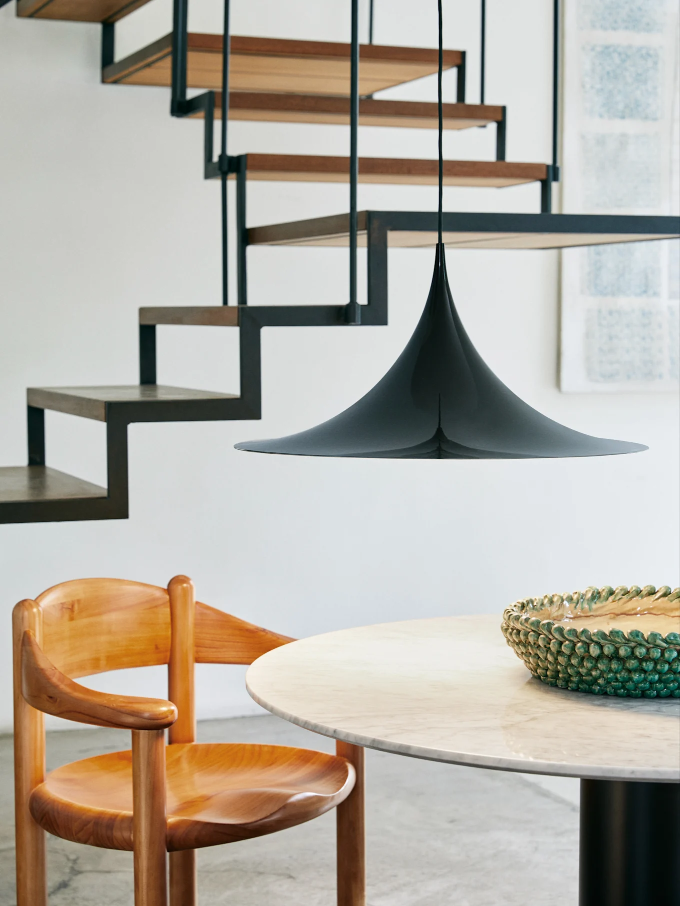 Une suspension noire conique est suspendue au-dessus d'une table ronde en marbre et d'une chaise en bois, avec un escalier moderne en arrière-plan.