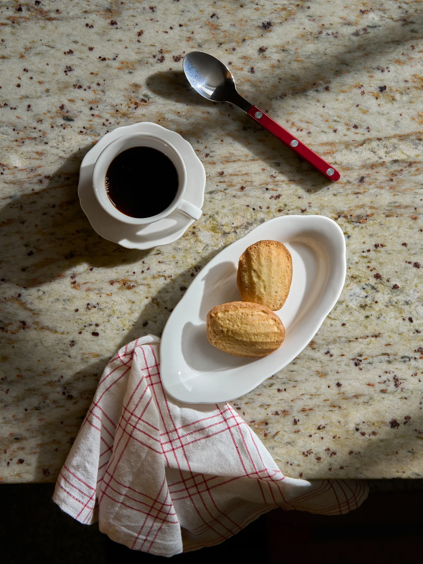 Vue de dessus: café noir en tasse, deux madeleines sur assiette, cuillère et serviette rayée sur comptoir en marbre.