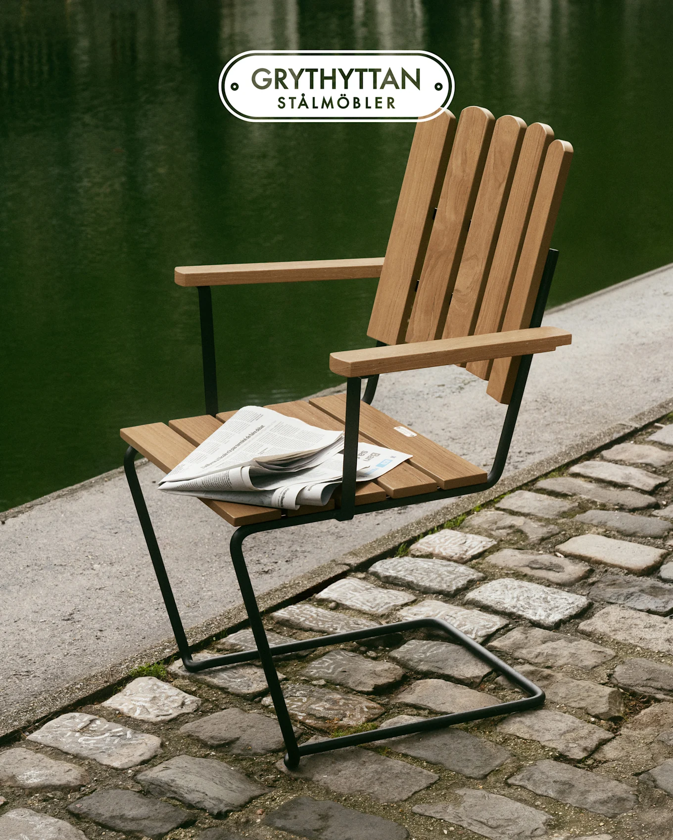 Chaise à lattes de bois avec cadre en métal noir sur un chemin pavé au bord de l'eau verte, journal sur l'assise.