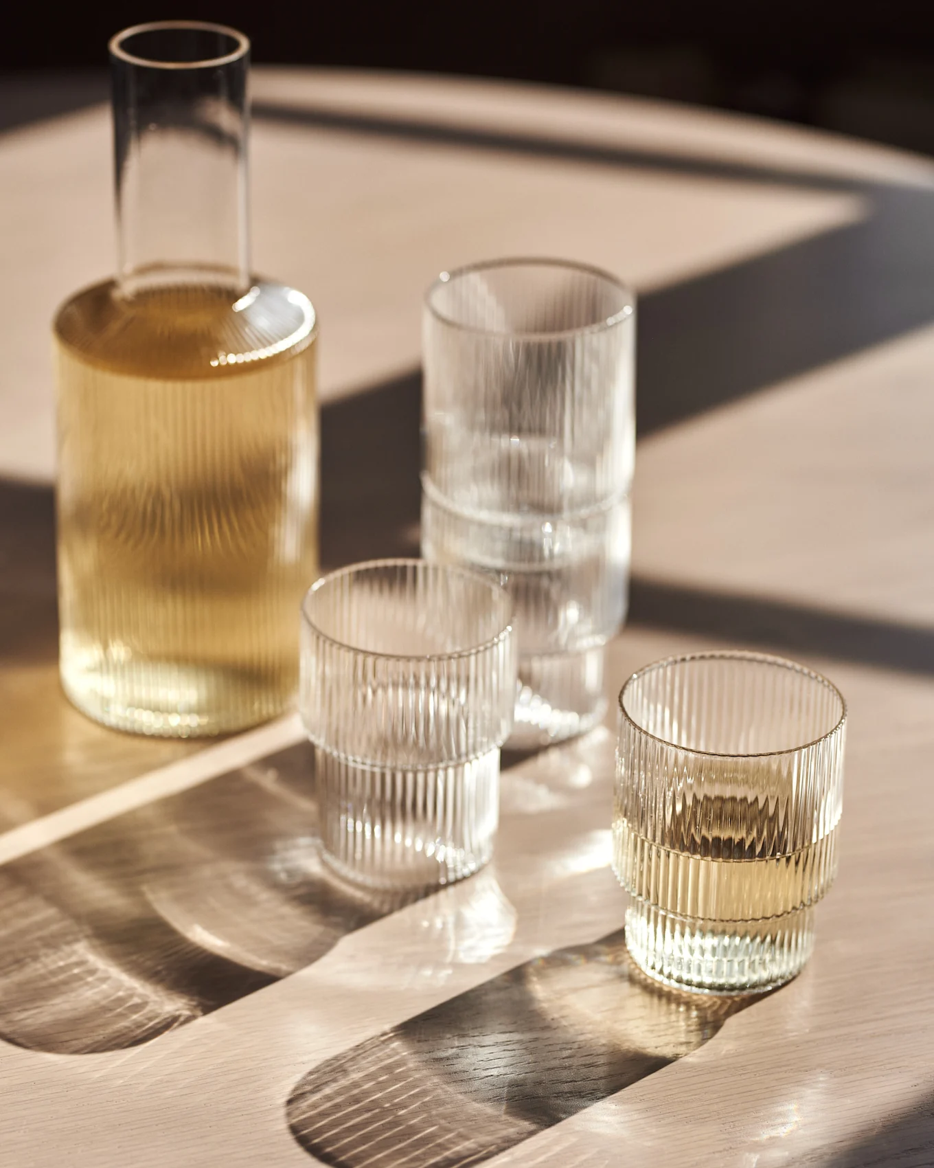 Carafe en verre et verres côtelés sur une table en bois sous la lumière du soleil.