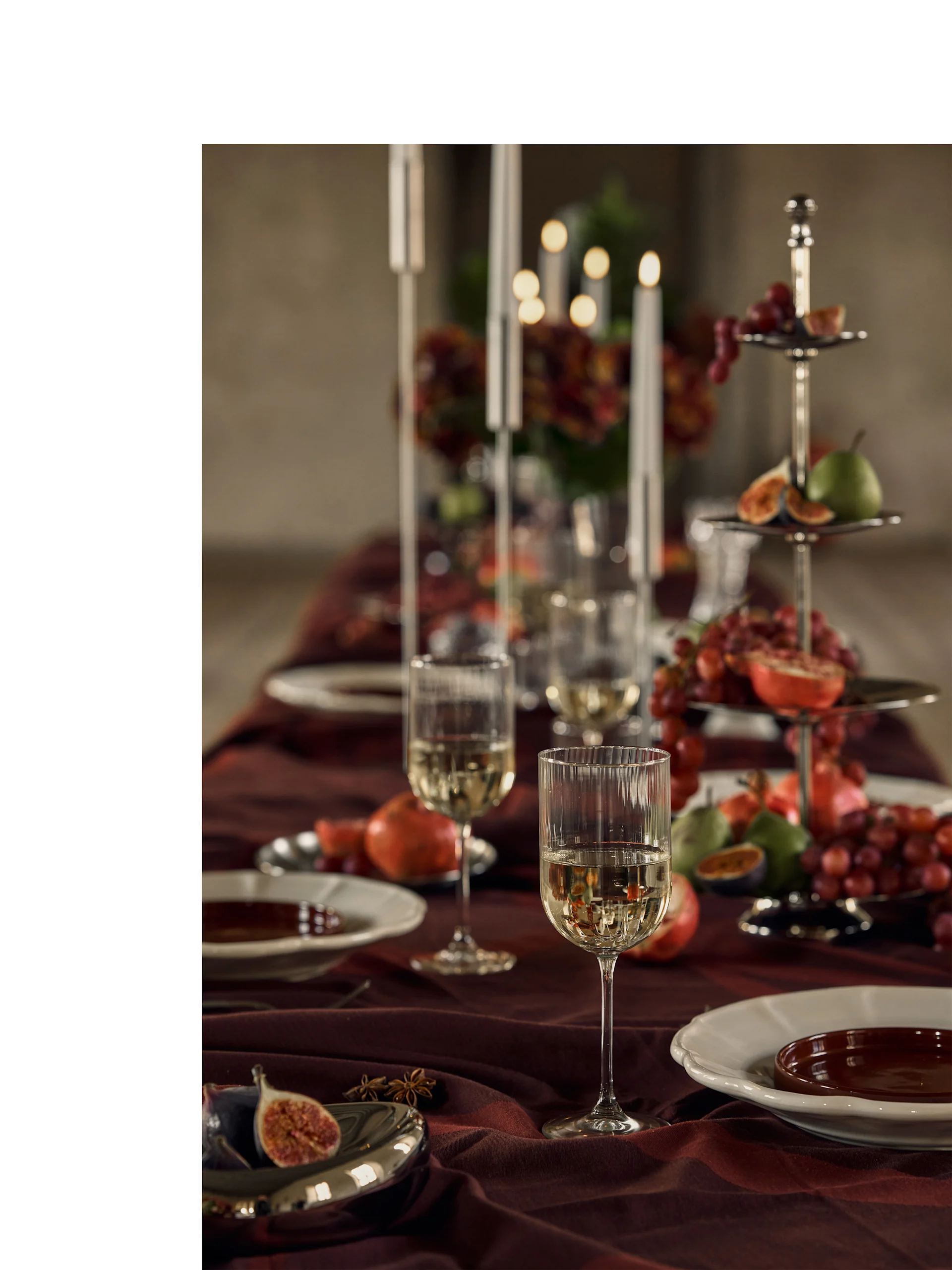 Table de fête dressée avec des verres de vin blanc, des assiettes et un support à étages rempli de fruits colorés et de bougies.