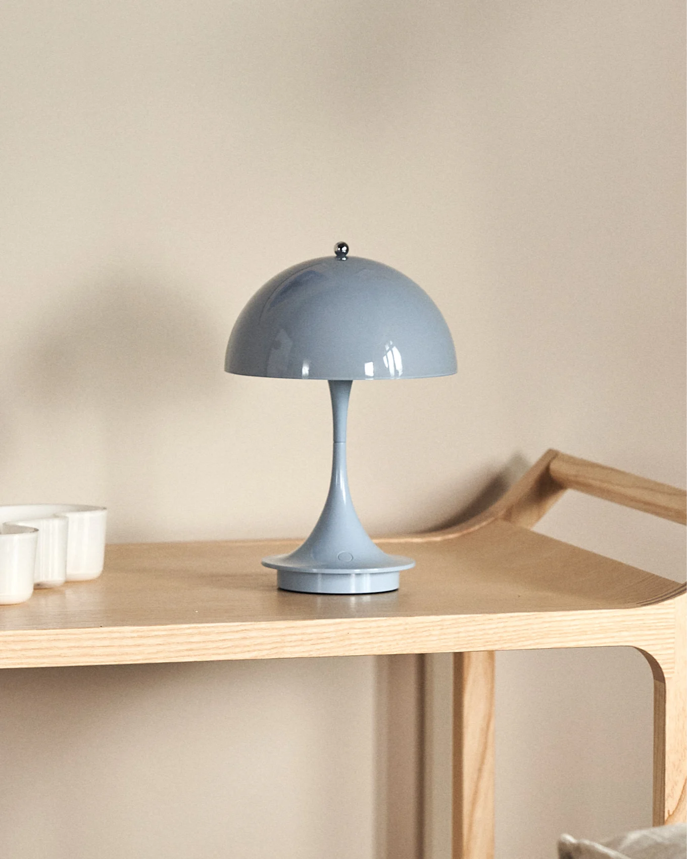 Une lampe de table bleu clair en forme de champignon sur une étagère en bois clair, avec des tasses blanches partiellement visibles à gauche.