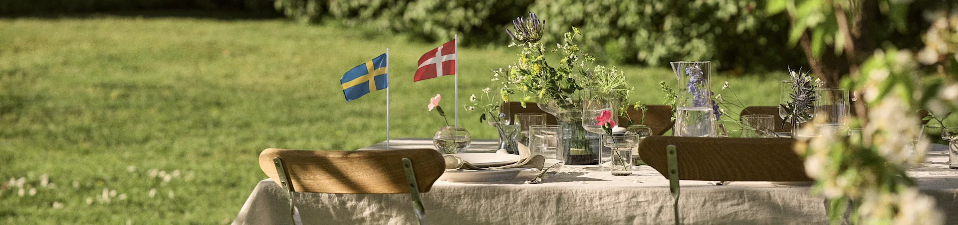 Table à manger d'extérieur dressée avec du linge blanc, de la verrerie, des arrangements floraux et des drapeaux suédois et danois dans un jardin ensoleillé.