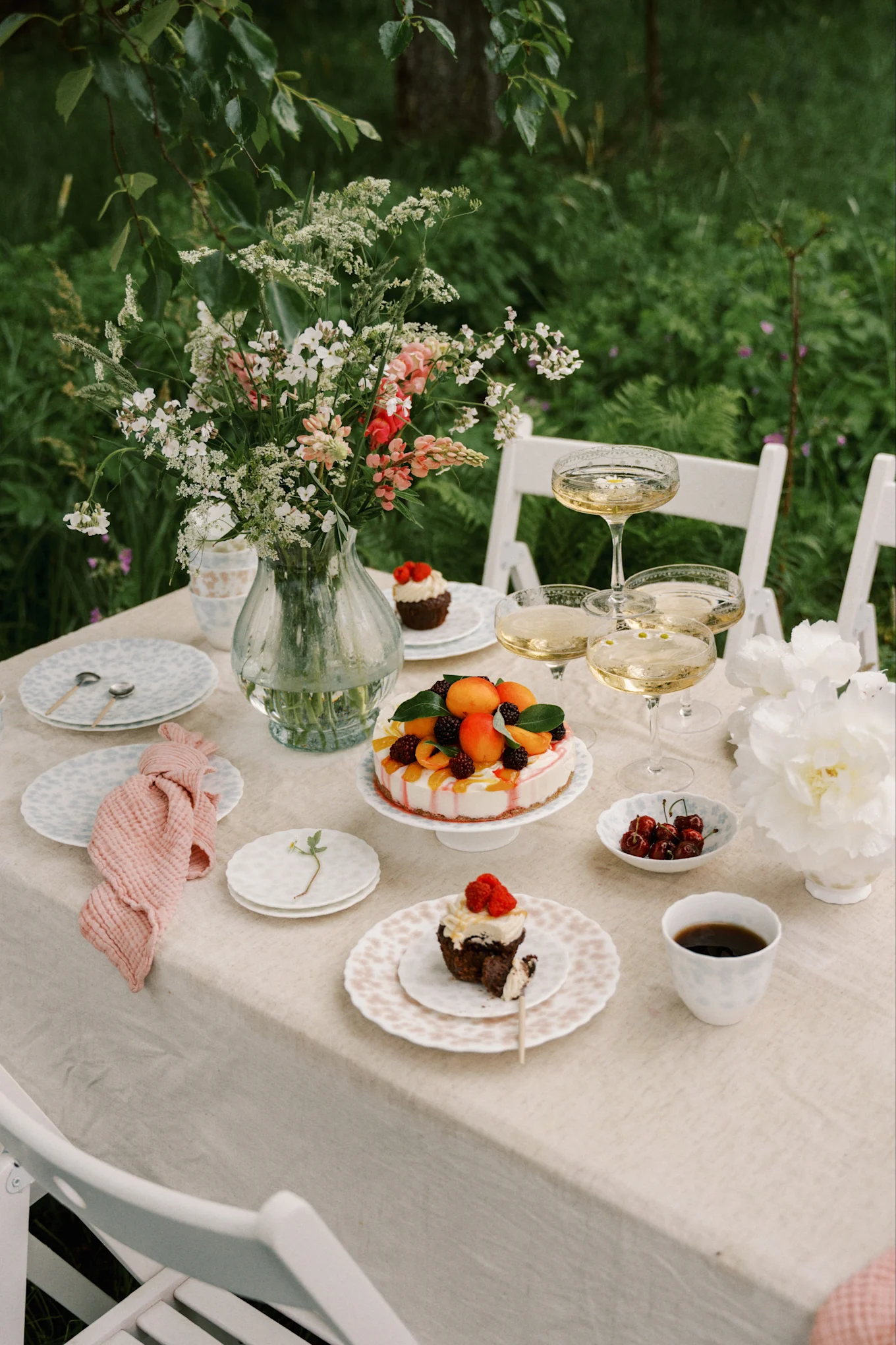Table d'extérieur dressée avec un vase de fleurs, du champagne, un gâteau aux fruits et des cupcakes aux baies.
