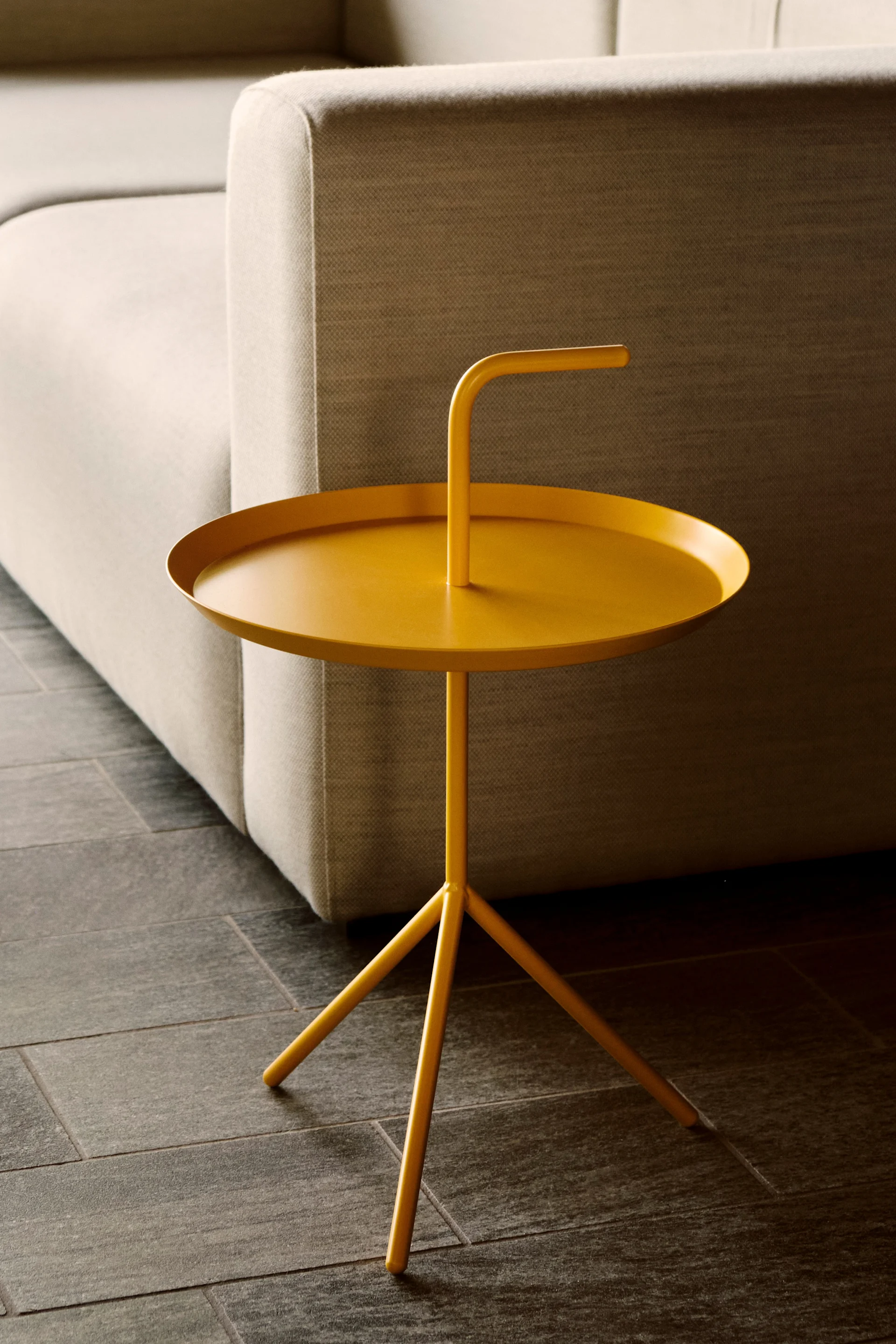 Une table d'appoint jaune vif, dotée d'un plateau rond et de trois pieds, est posée à côté d'un canapé clair sur un sol en carrelage foncé.