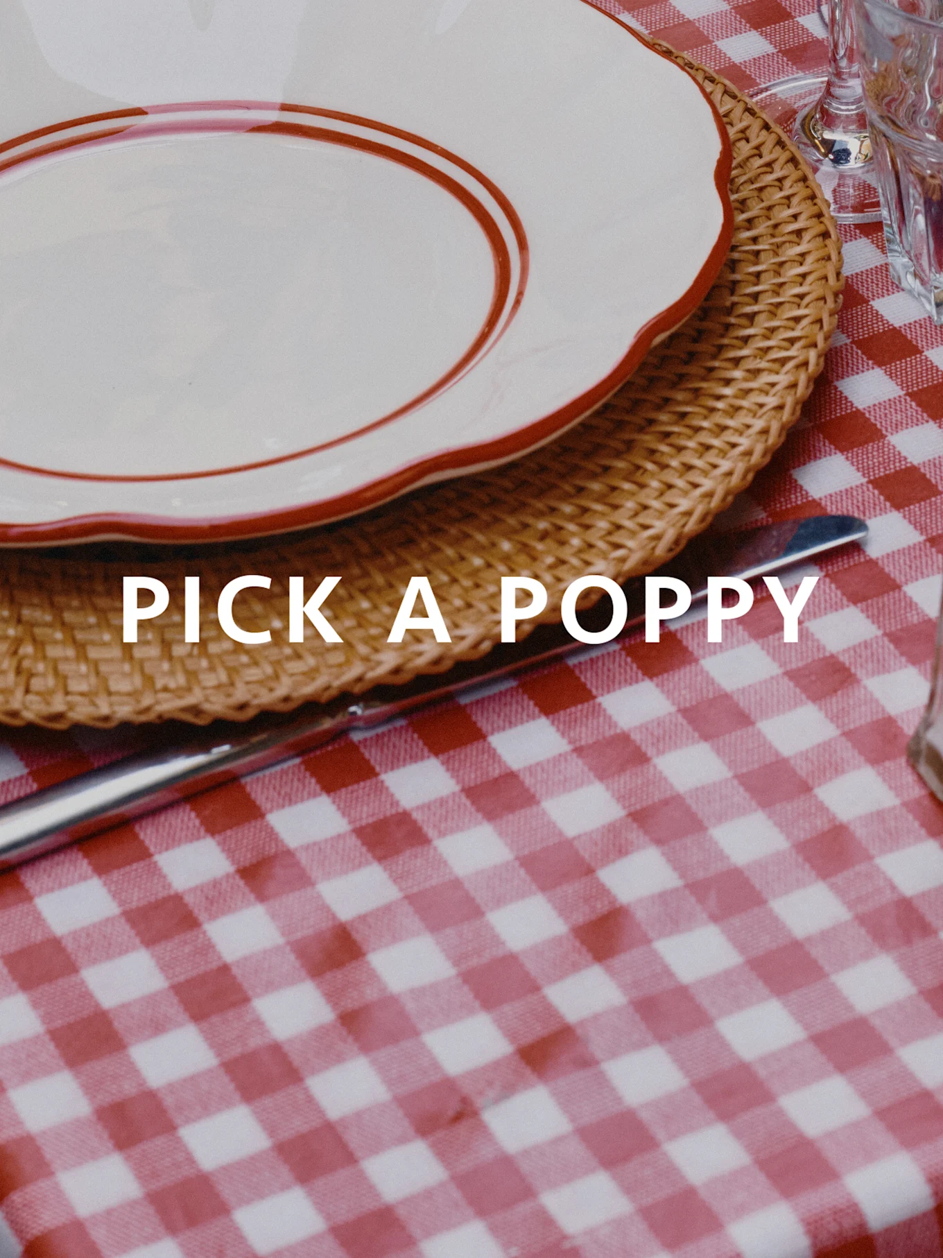 Table dressée avec nappe à carreaux rouge et blanc, set de table en osier et assiette blanche à bord rouge. Texte : PICK A POPPY.