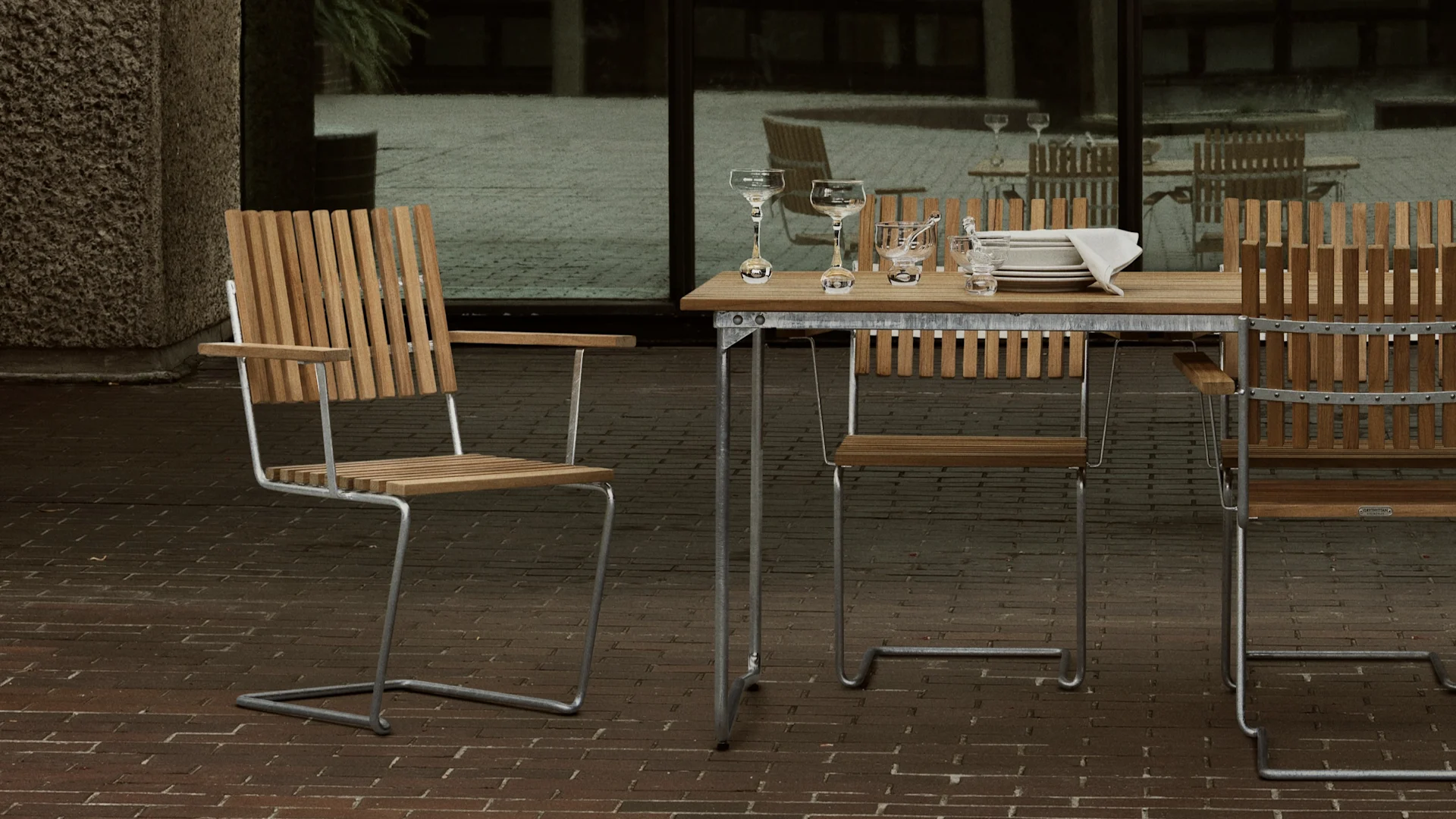 Ensemble de salle à manger d'extérieur avec table et chaises en bois sur une terrasse en briques, dressé avec verrerie et assiettes.