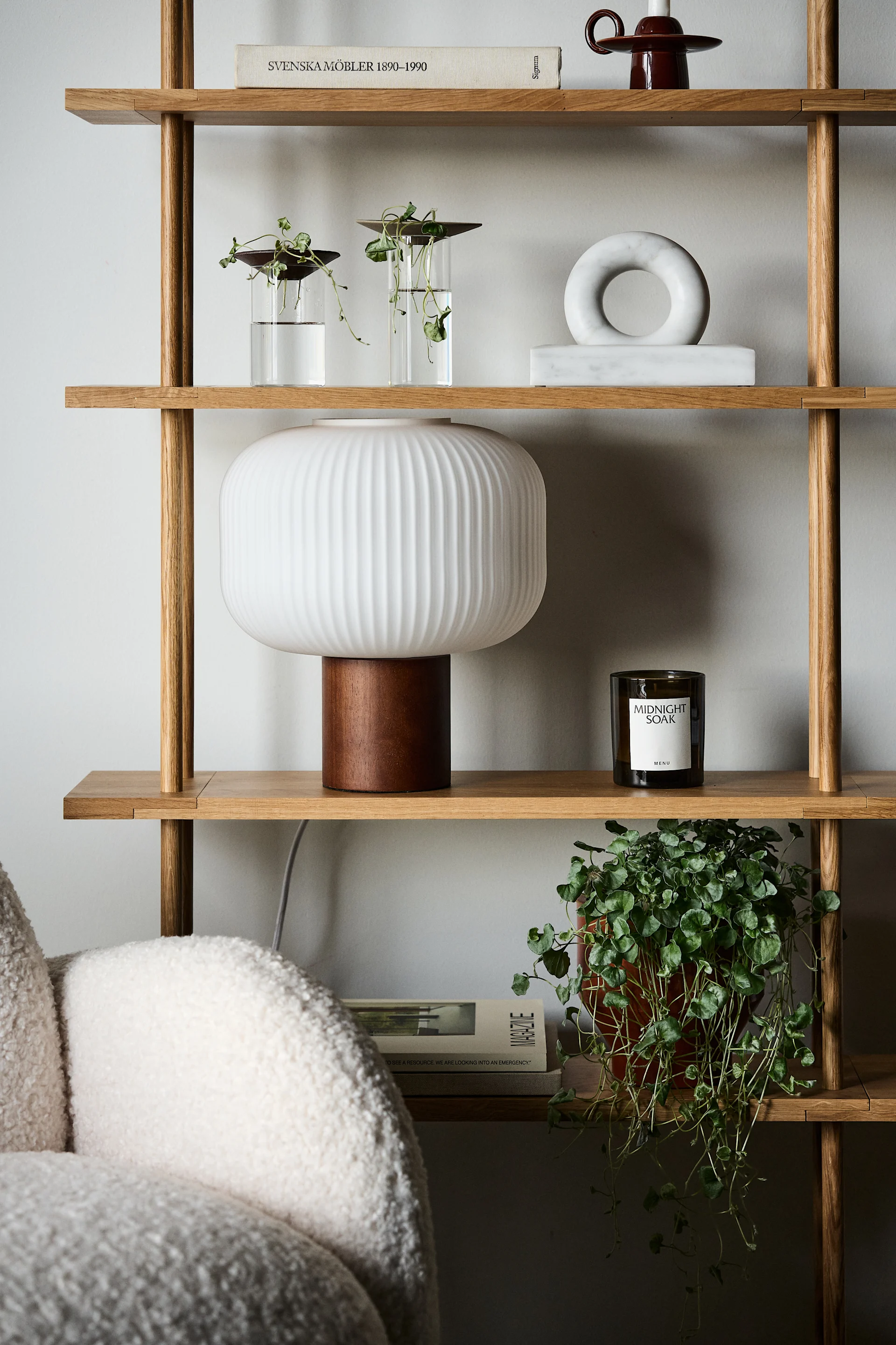 Une étagère en bois clair avec des plantes, une lampe blanche cannelée, des décors en marbre, un livre et une bougie.