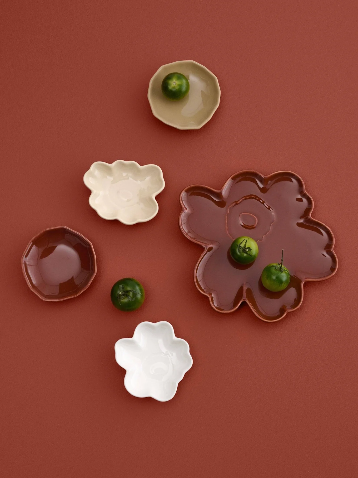 Assortiment de petites assiettes de formes irrégulières en crème, blanc et marron avec de petits fruits verts, sur un fond brun-rouge.