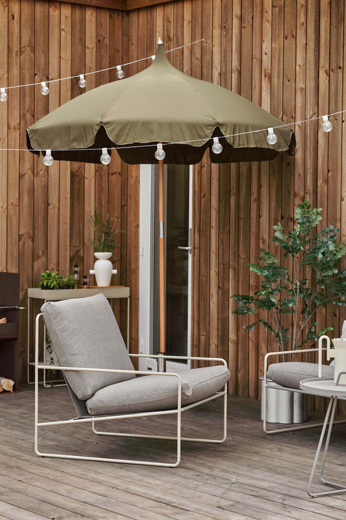 Une terrasse en bois accueillante avec des lumières féeriques, un parasol Lull et une chaise longue Desert, tous deux de Ferm Living.