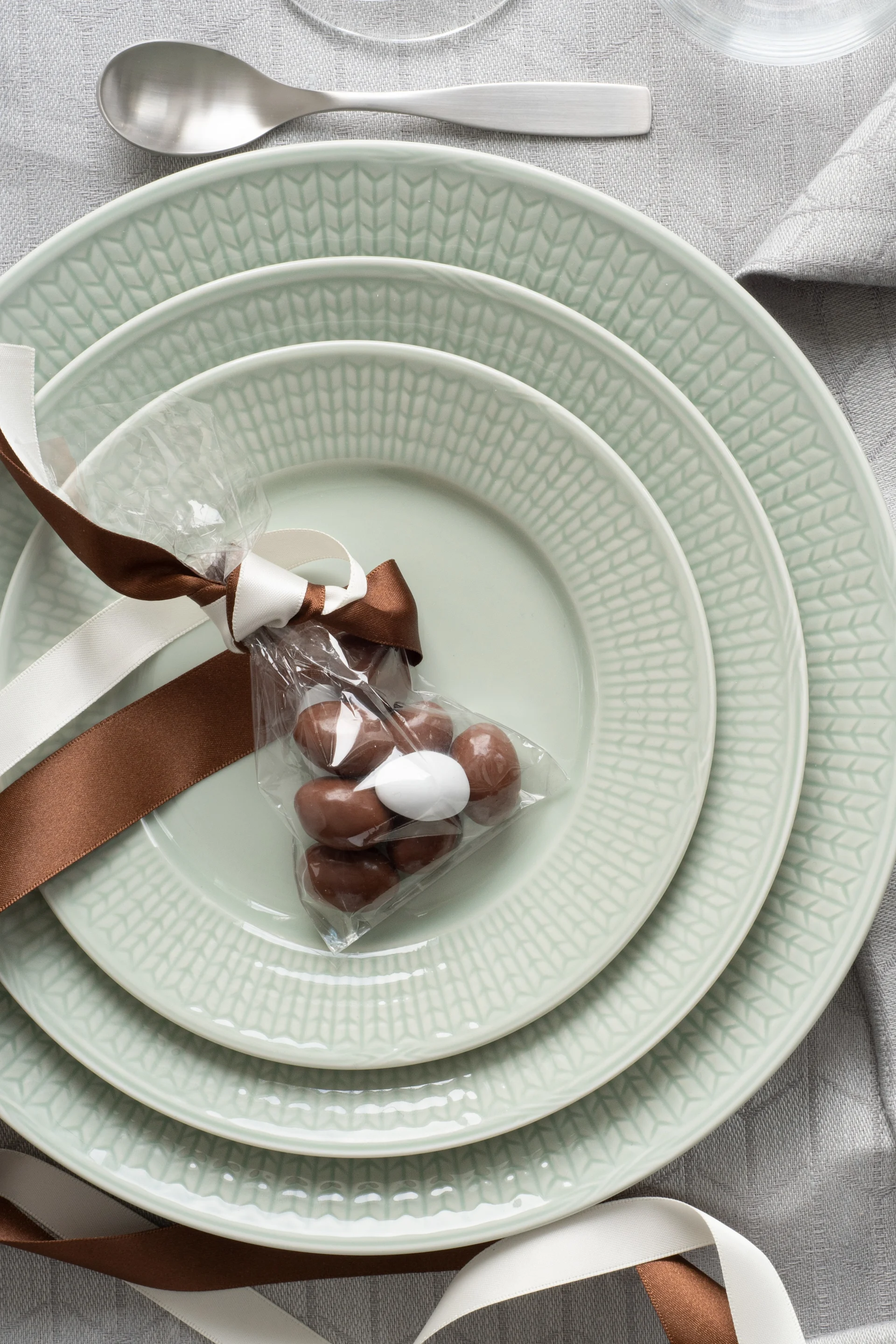 Assiettes empilées vert clair avec un sac cadeau de chocolats sur une nappe grise, à côté de couverts argentés.