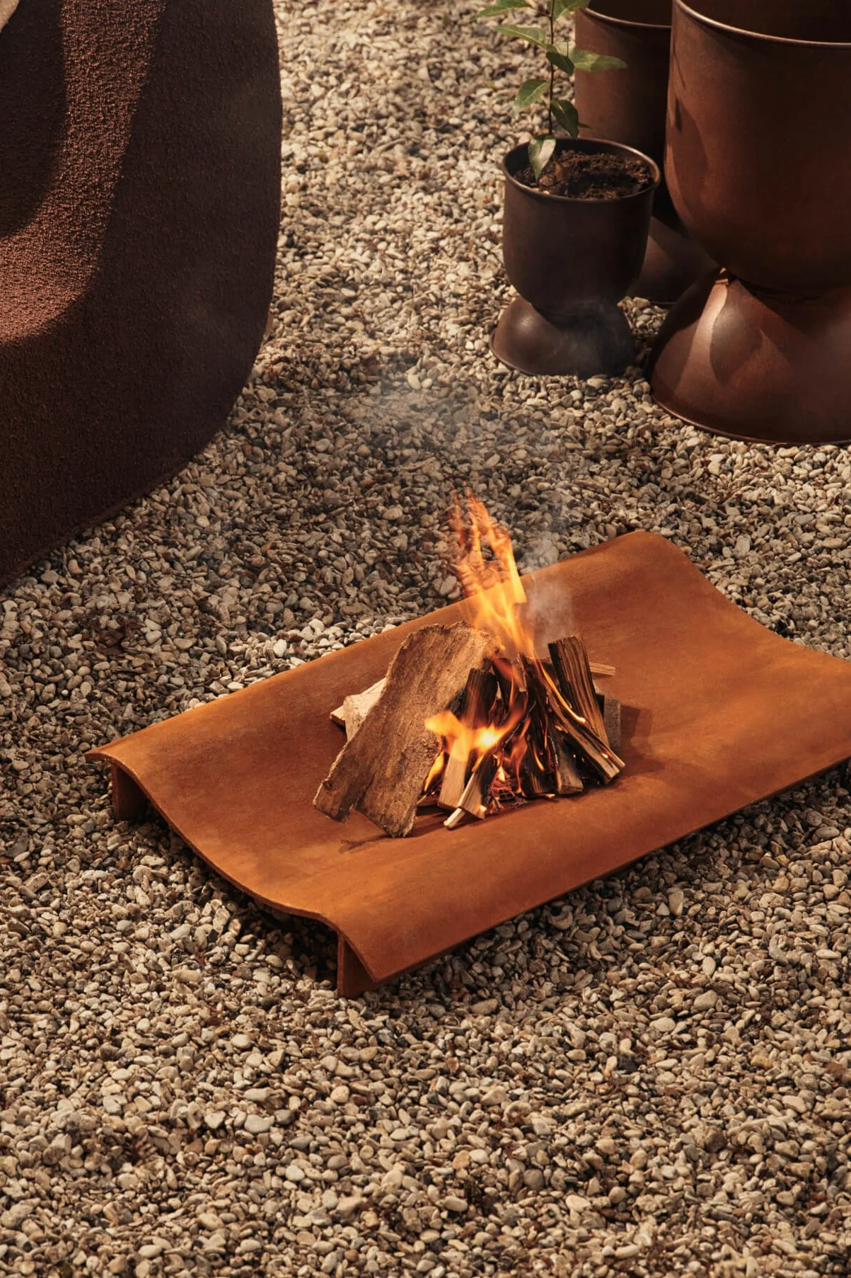 Un petit feu de camp brûle sur un plateau métallique incurvé, de couleur orange rouillé, posé sur un sol recouvert de gravier.