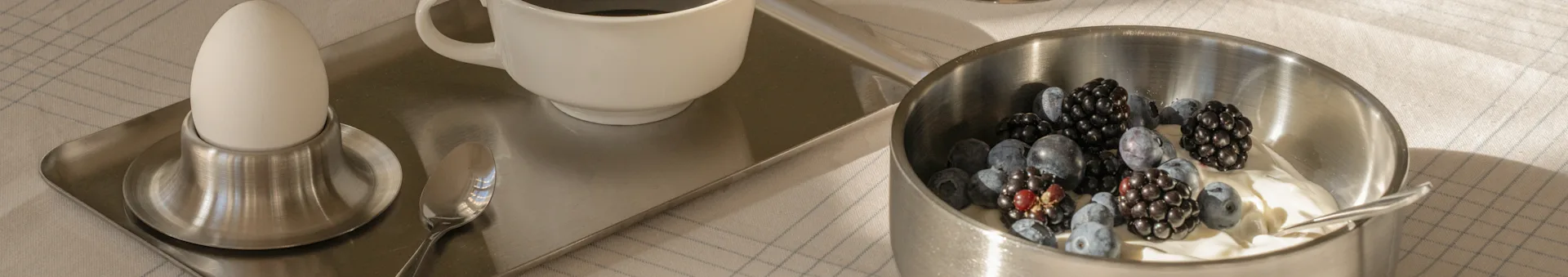 Plateau de petit-déjeuner avec un œuf dur, du café et un bol de yaourt garni de myrtilles et de mûres sur une nappe à carreaux.