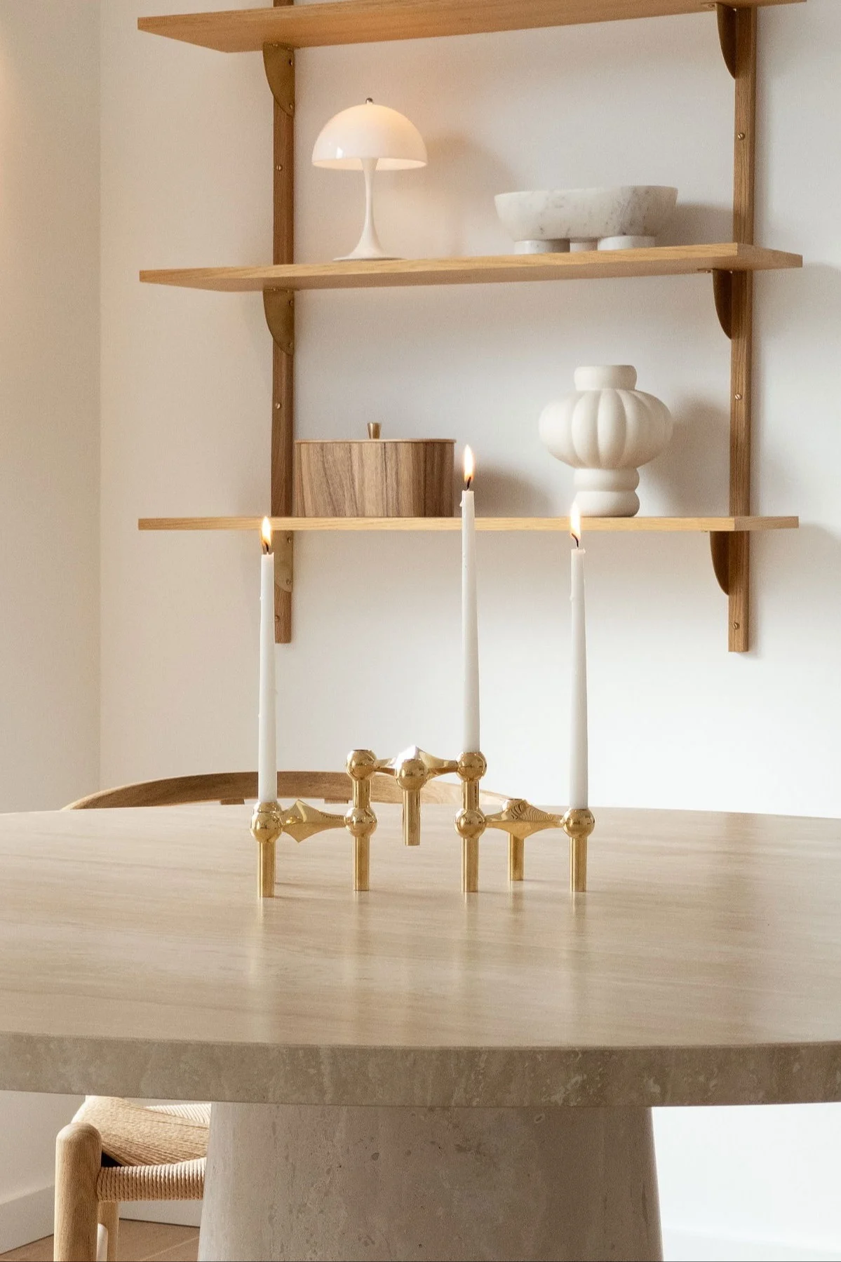 Un candélabre doré surmonté de trois bougies allumées, posé sur une table ronde, avec derrière des étagères sur lesquelles sont disposés des objets décoratifs.