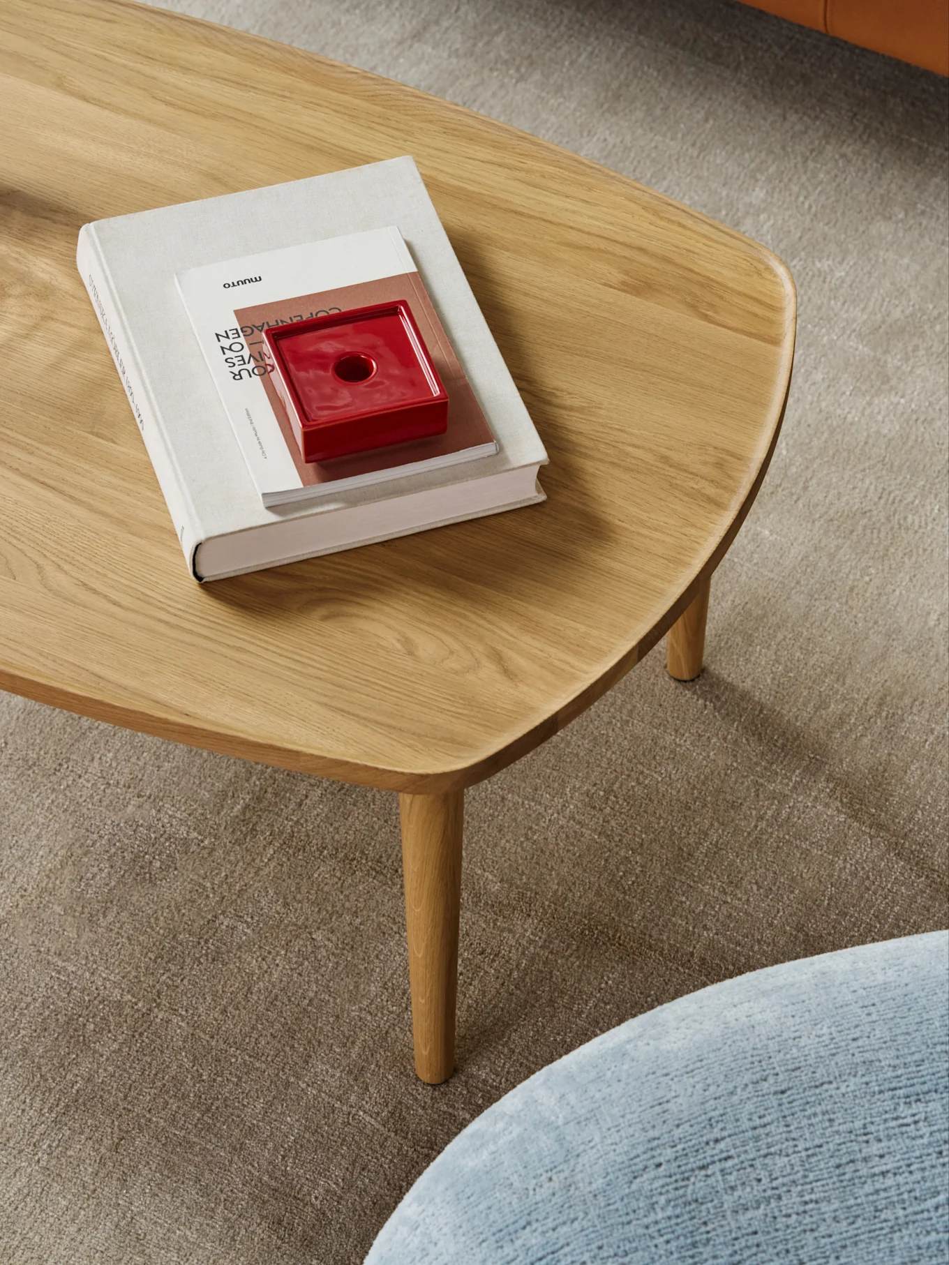 Une table basse en bois clair aux bords arrondis, avec une pile de livres et un objet en céramique rouge, sur un tapis beige.