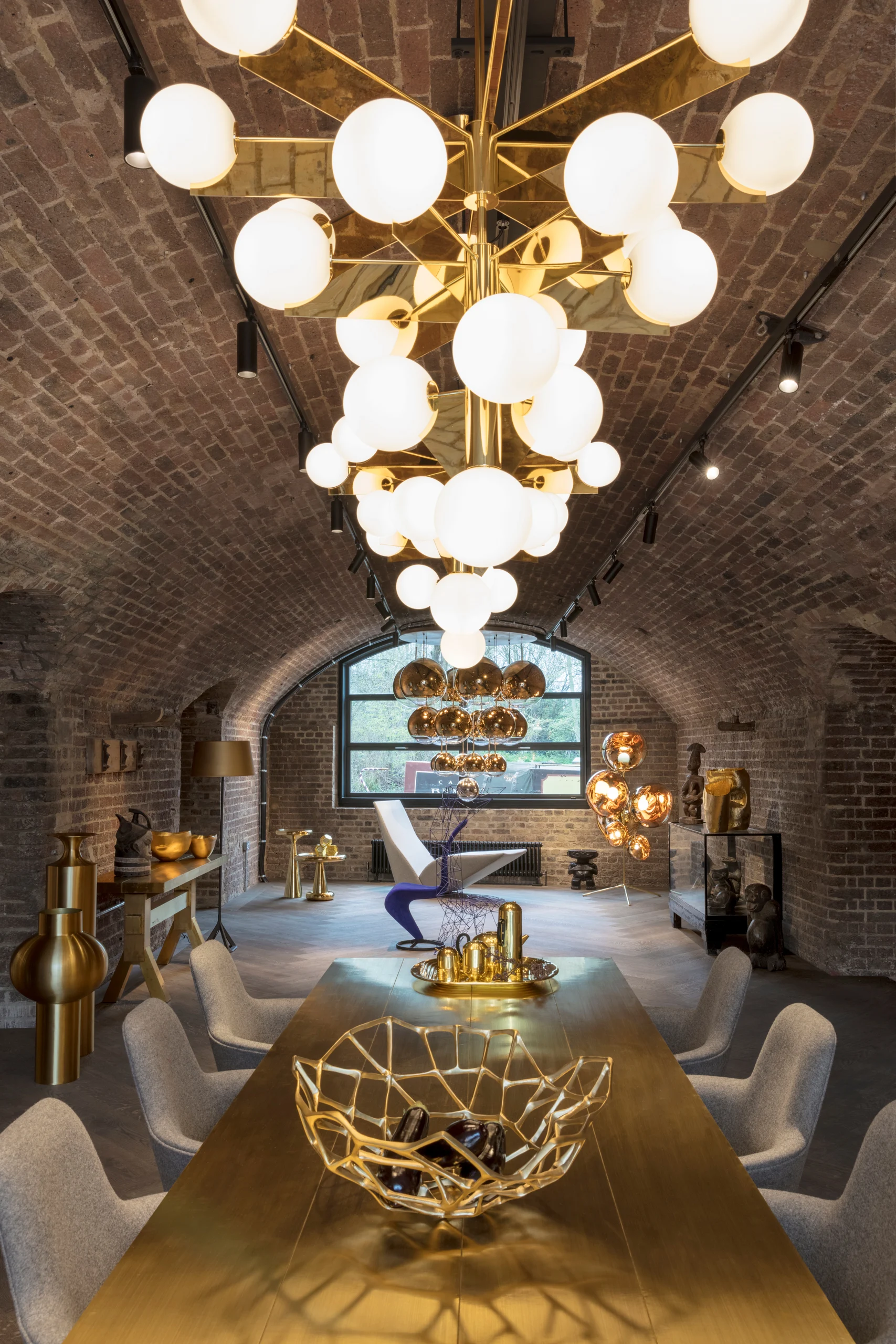 Longue table dorée avec un bol géométrique et des chaises grises sous un lustre doré étagé dans une salle voûtée en briques.